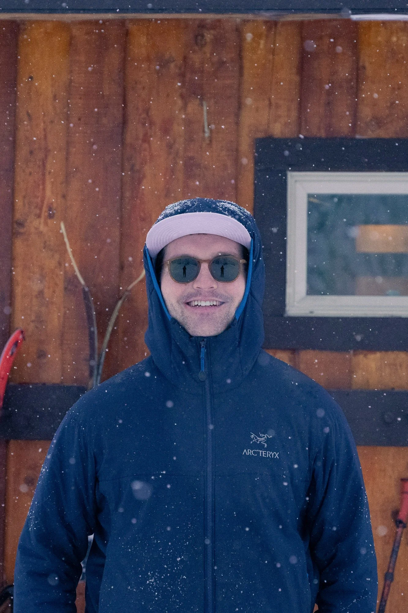Man smiling in snowy outdoor setting, wearing sunglasses, a blue Arc'teryx jacket, and a cap, with a wooden wall and window in the background.