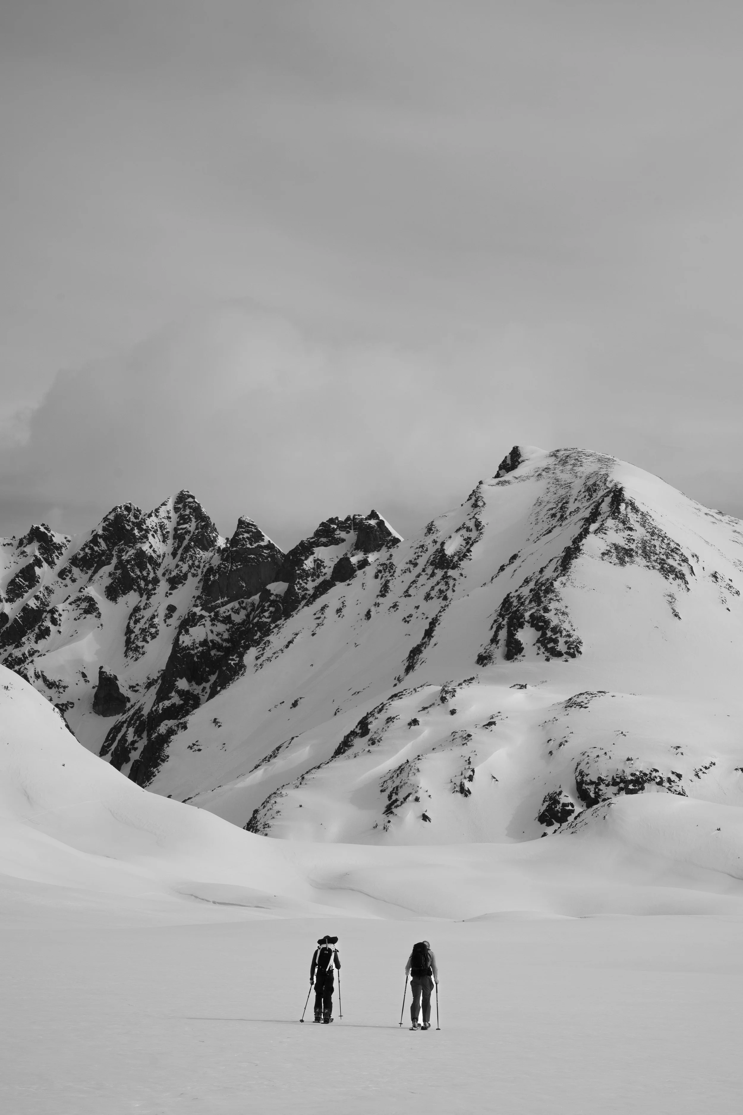 Two hikers with backpacks and trekking poles walking across snow-covered landscape in front of snow-capped mountains under cloudy sky.