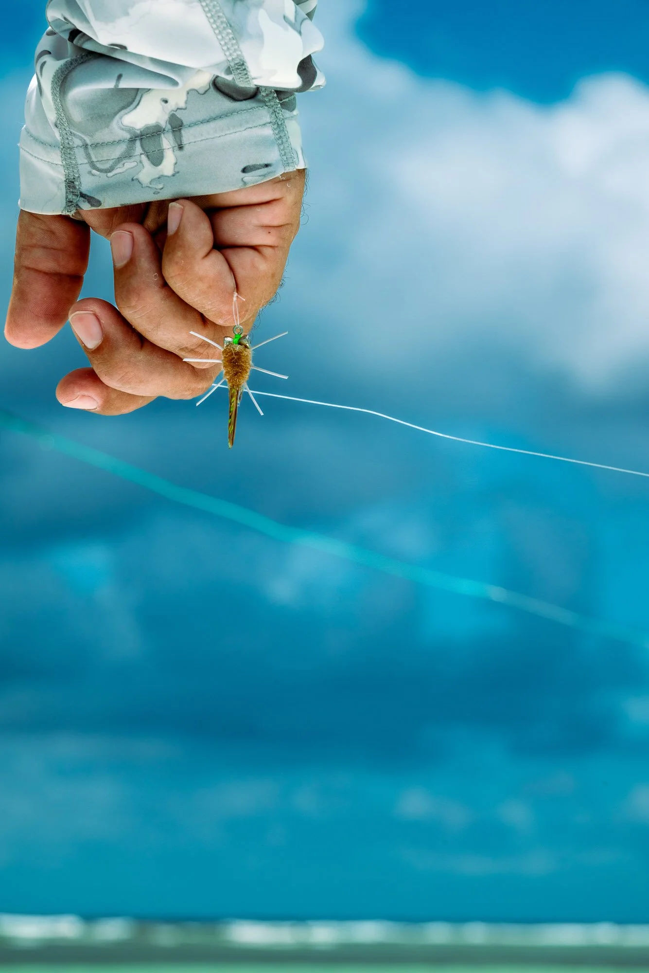fly fishing. A person's hand holding a fishing lure that resembles a small fish with a fuzzy body and long tail, with a cloudy sky in the background. Turneffe Flats, Belize