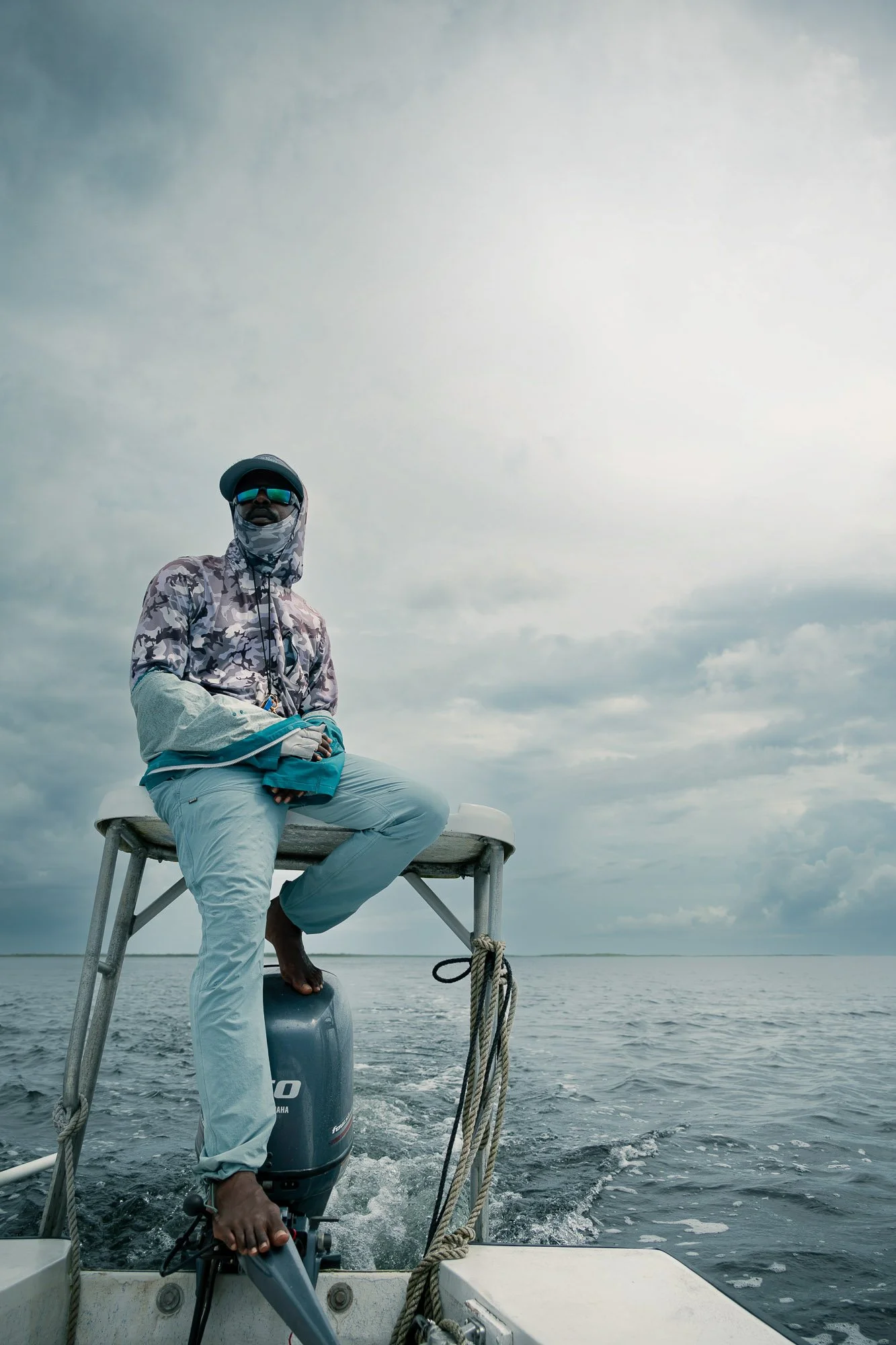 Permit fly fishing Guide sitting on the edge of a boat with an outboard motor, on open water with cloudy skies. Turneffe Flats, Belize