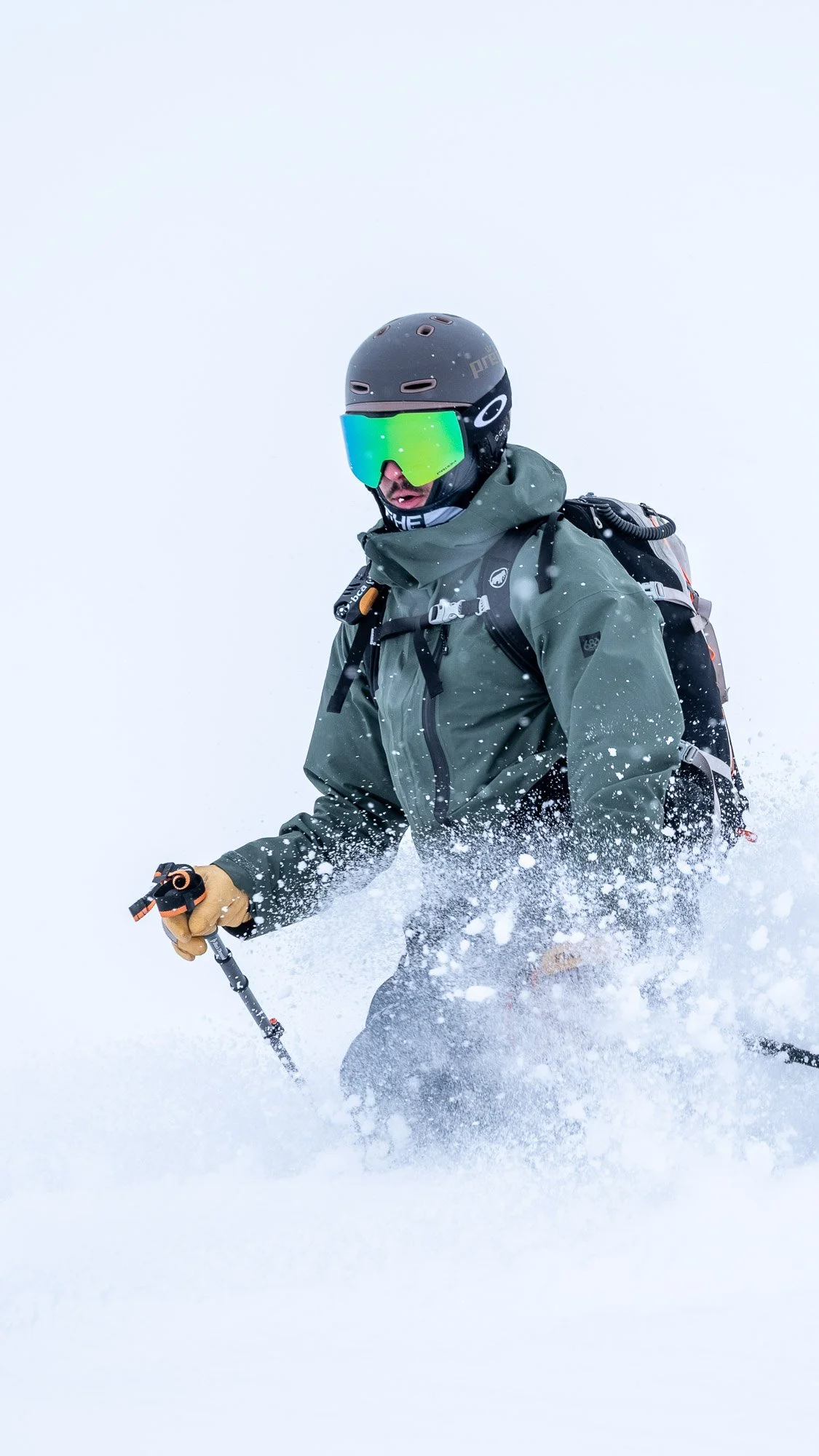 A skier wearing a black helmet, green goggles, gray jacket, tan gloves, and carrying a backpack is skiing through snow in a cloudy, snowy environment.