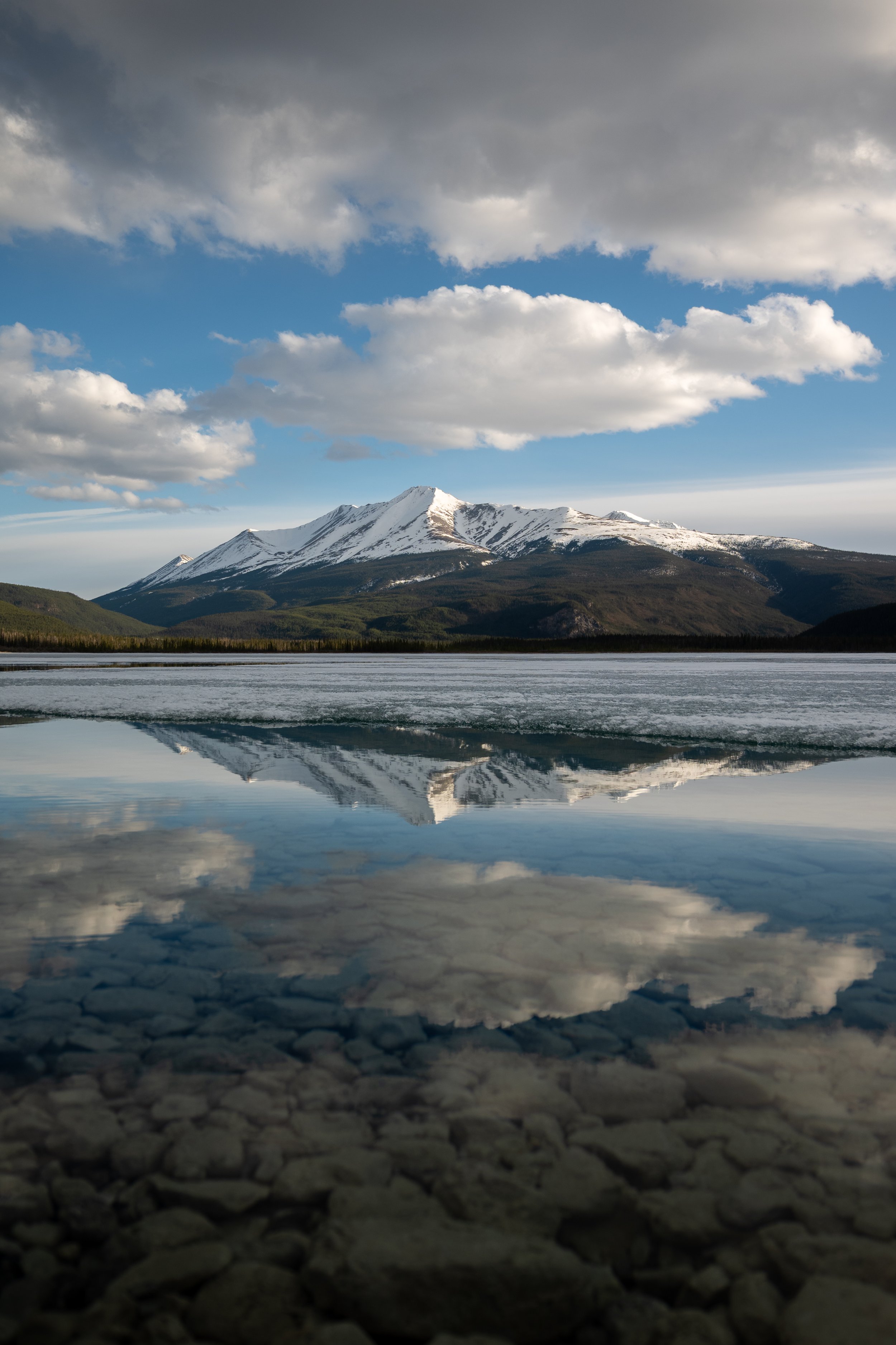Snow-capped mountain reflected in a partially frozen lake with a rocky bottom and a cloudy sky.
