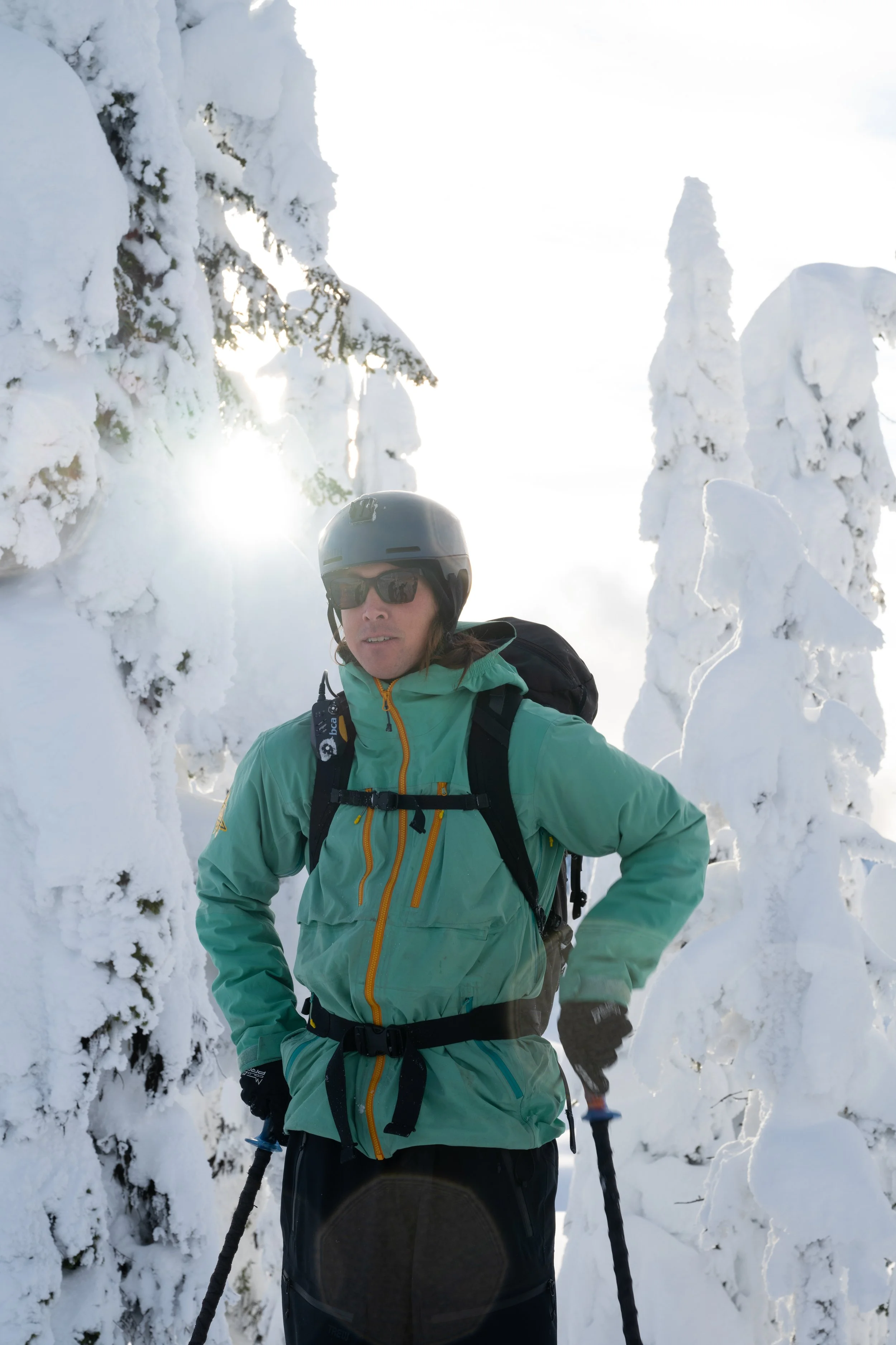 A man in a green winter jacket, black helmet, and sunglasses, carrying a backpack, standing in a snow-covered forest with snow-laden trees.