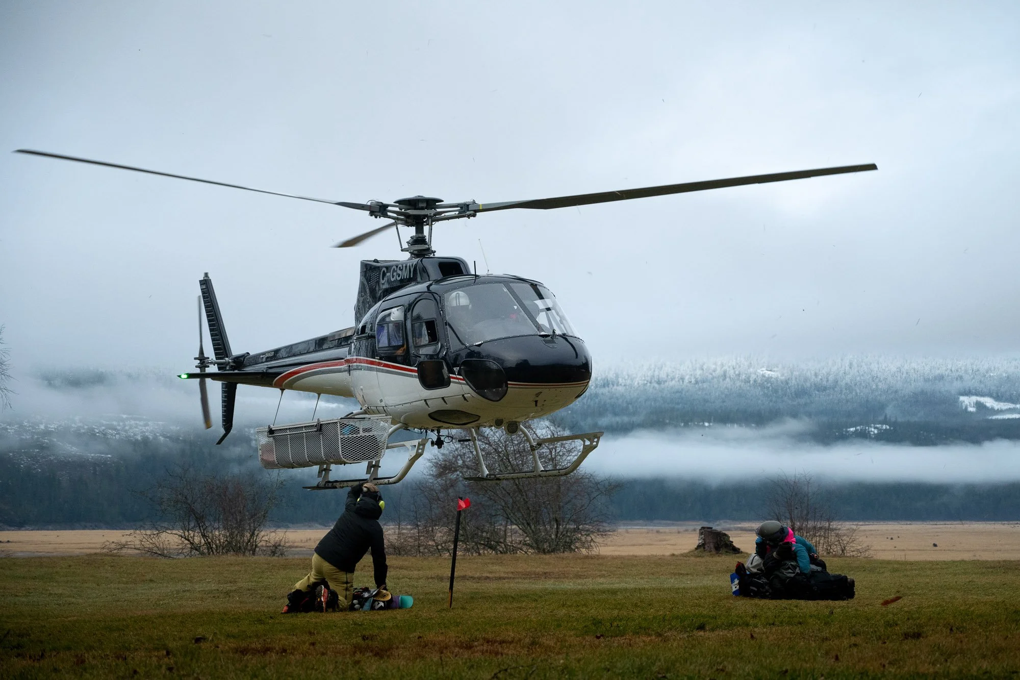 A helicopter hovering above the ground while two people prepare or pack equipment on a grassy field. Revelstoke, British Columbia Canada