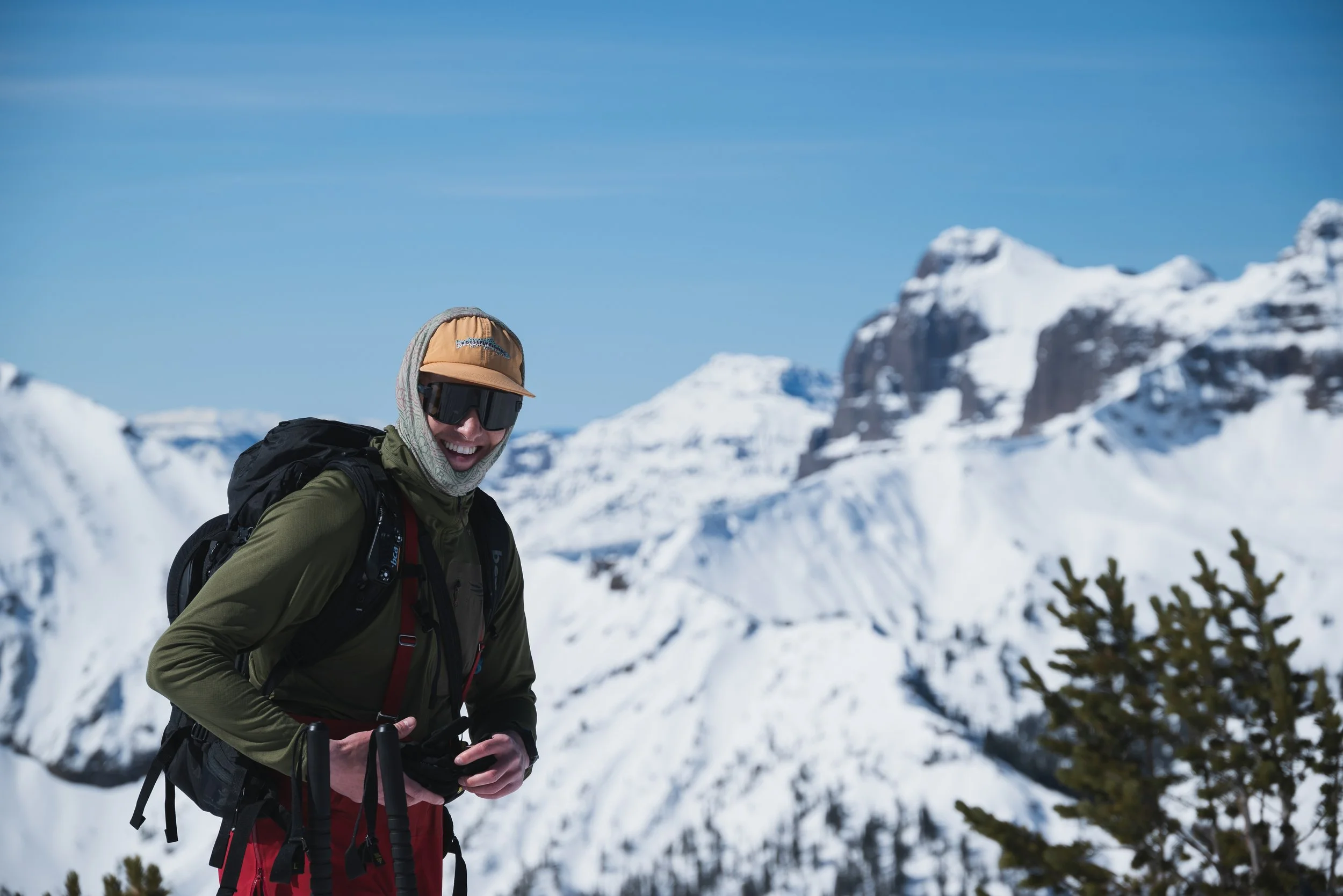 A smiling person dressed in winter outdoor gear, including a green jacket, red pants, a beige cap, and sunglasses, standing in a snowy mountain landscape with snow-covered peaks and a clear blue sky.