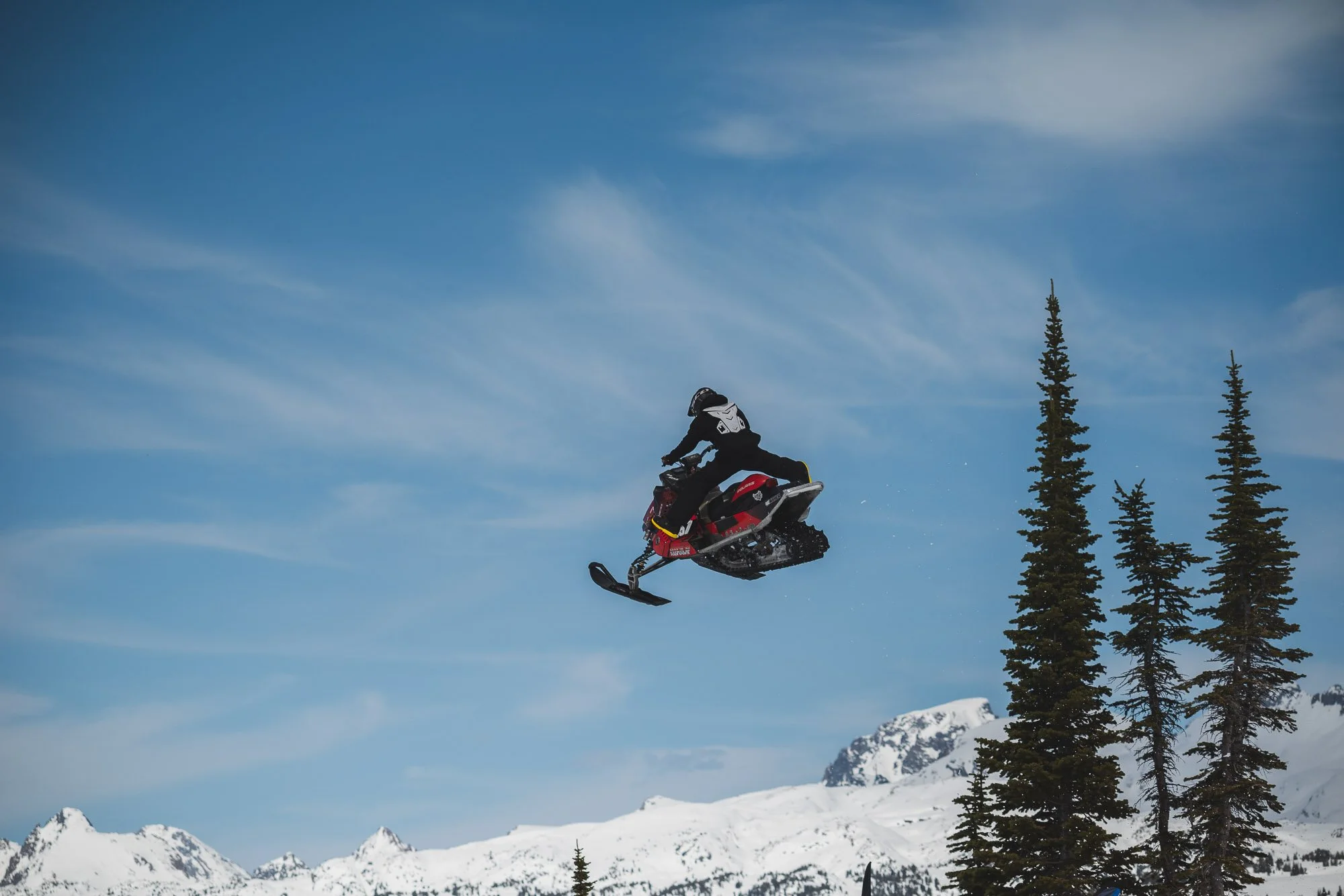 Person riding a snowmobile mid-air over snowy mountains and pine trees on a clear, blue day. Montana backcountry snowmobiling