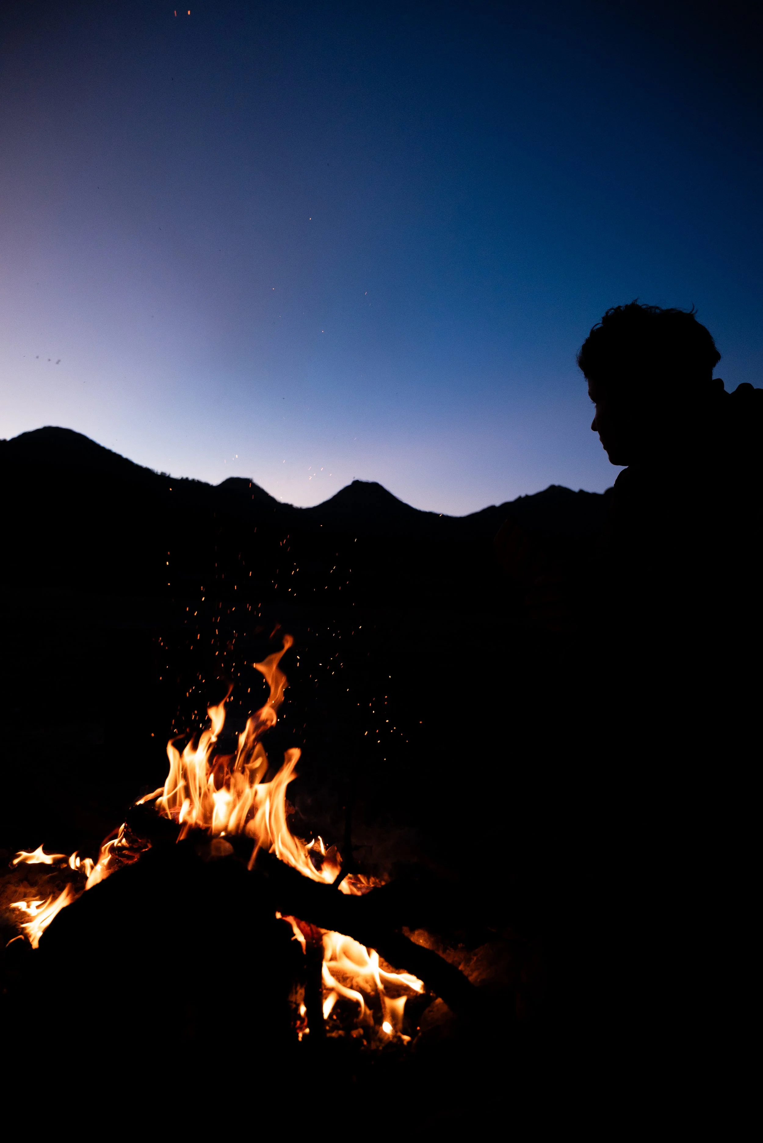 Silhouette of a person sitting by a campfire in a mountainous landscape during twilight, with a clear sky and visible stars.