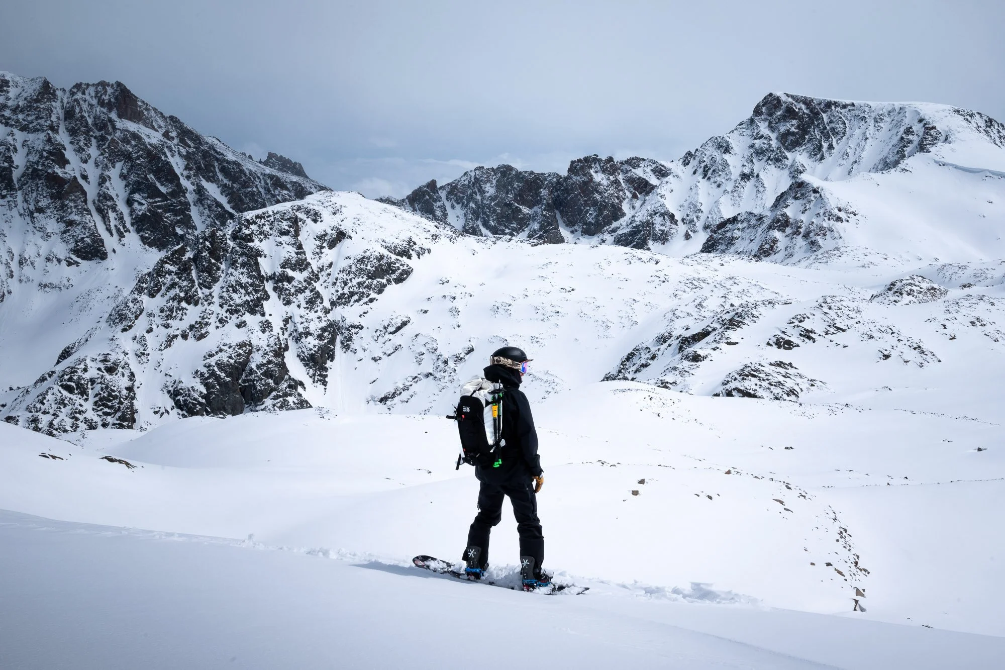 A person in winter gear with a backpack snowboarding on a snowy mountain landscape with rocky peaks in the background. Montana Backcountry skiing
