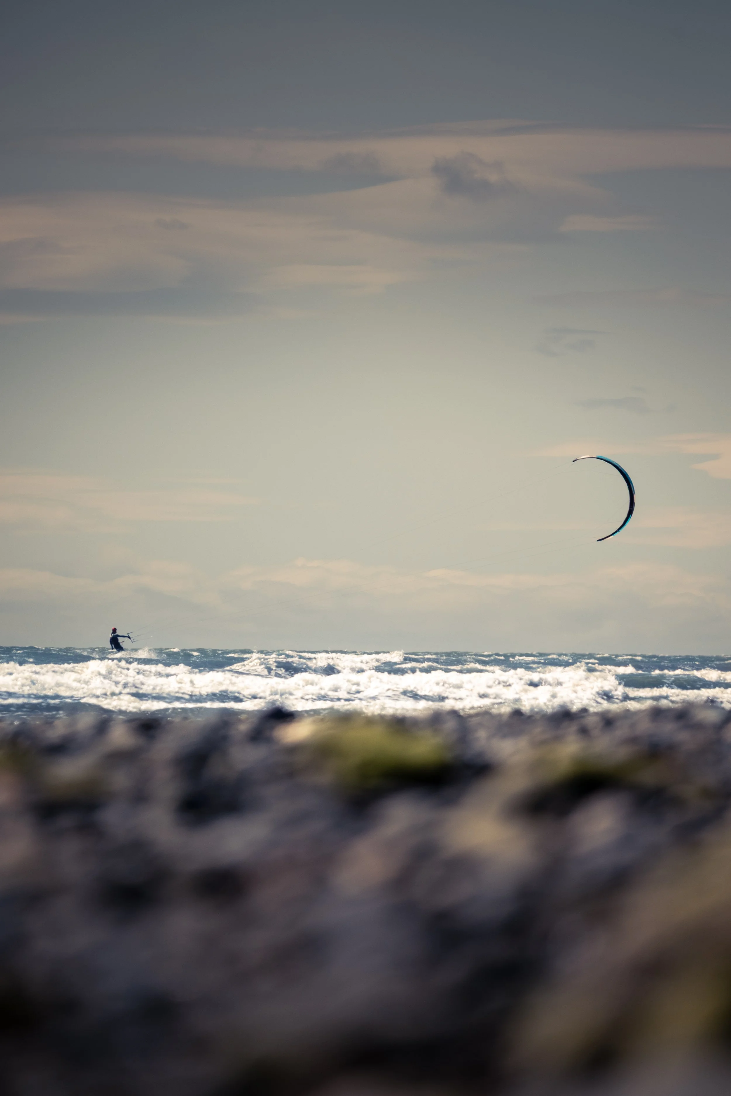 A person kiteboarding on the ocean with a crescent-shaped kite in the sky, with a blurred foreground of rocks or seaweed.