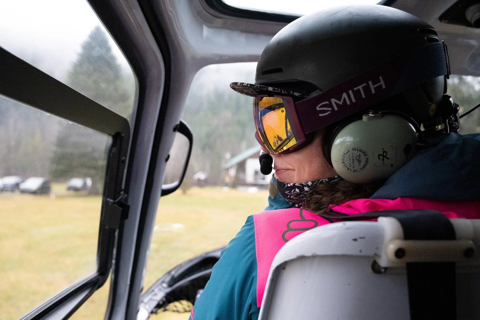 Person wearing a helmet, goggles, and a pink vest, sitting in the cockpit of a helicopter, looking out the window at the cloudy landscape outside.