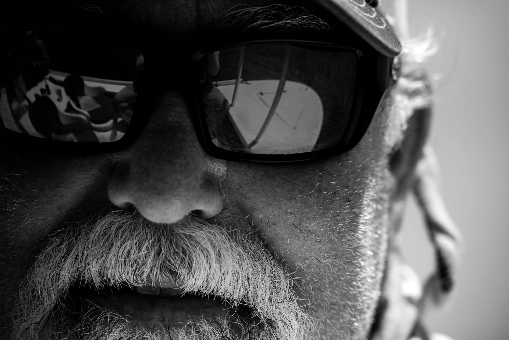Close-up black-and-white photo of an older man with a beard and mustache. He is wearing dark sunglasses that reflect a scene with multiple people and glasses. The man's face is detailed with textured skin and facial hair.