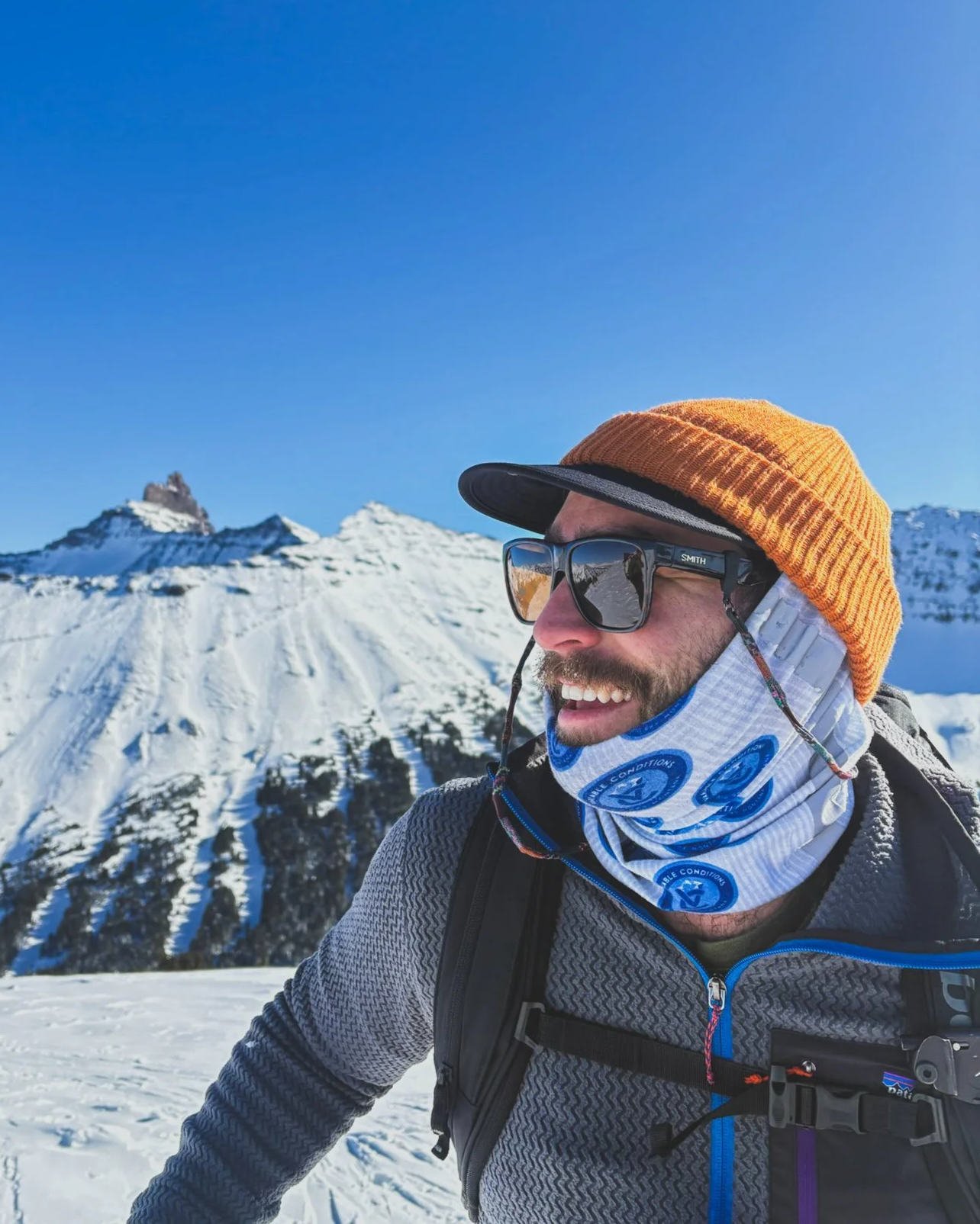 Man wearing a tan knit beanie, sunglasses, and a neck gaiter with blue logos, smiling in snowy mountain landscape.