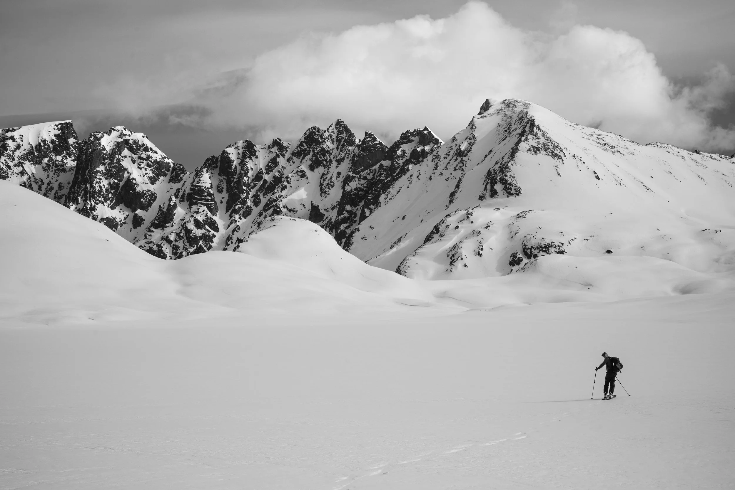 A person skiing across a snowy, flat landscape with snow-covered mountains and cloudy sky in the background, black and white photograph.