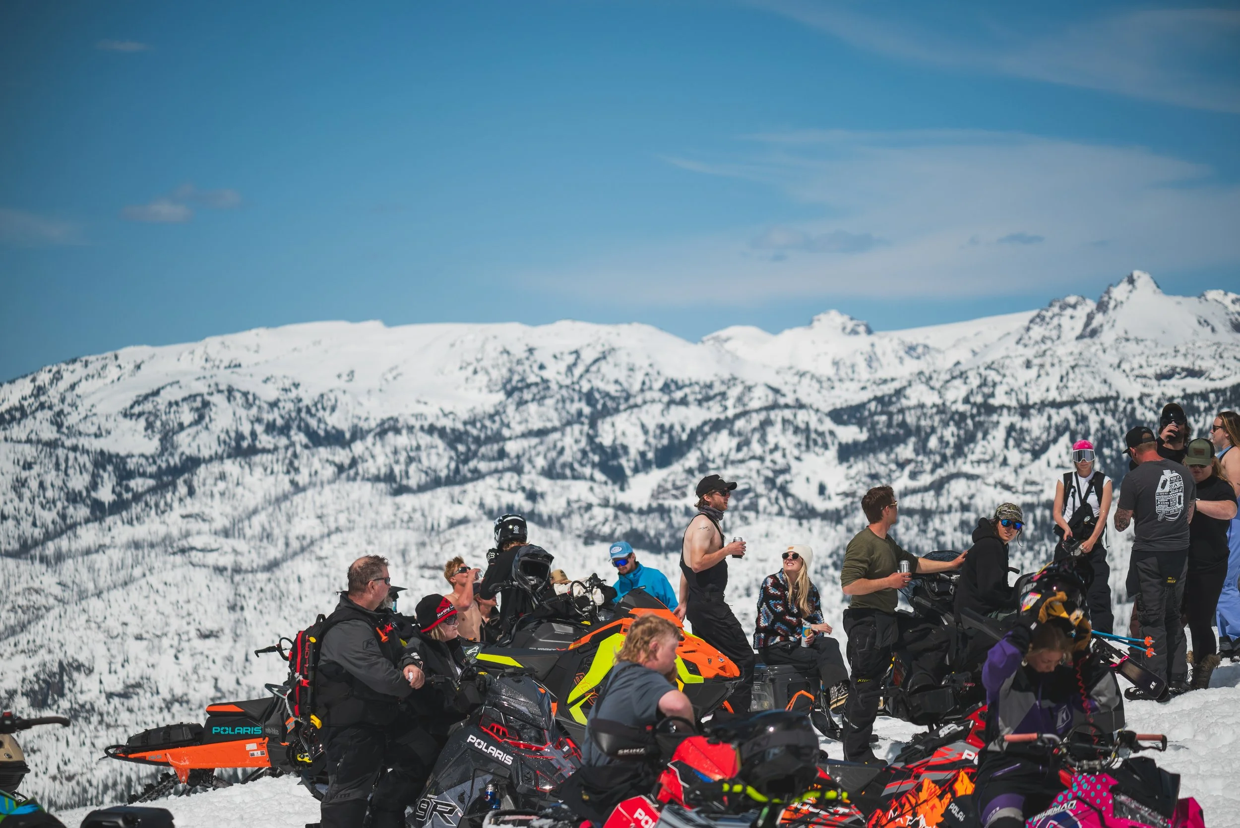 Group of people on snowmobiles and standing in a snowy mountain landscape with snow-covered peaks and a blue sky.