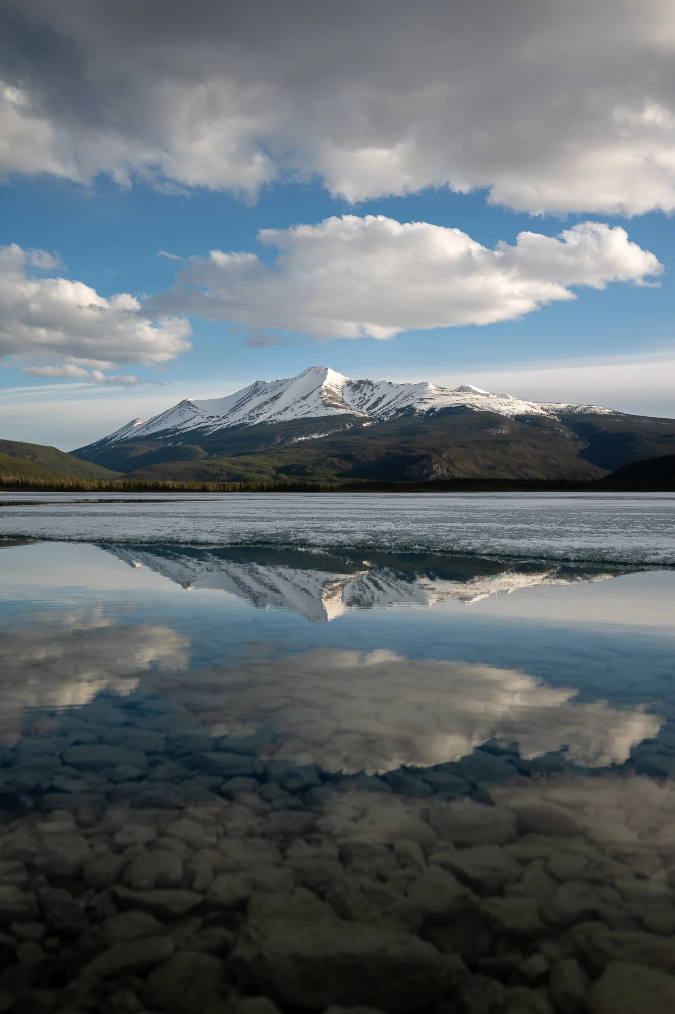 Snow-capped mountain reflected in a calm lake with a cloudy sky overhead. Muncho Lake - British Columbia, Canada