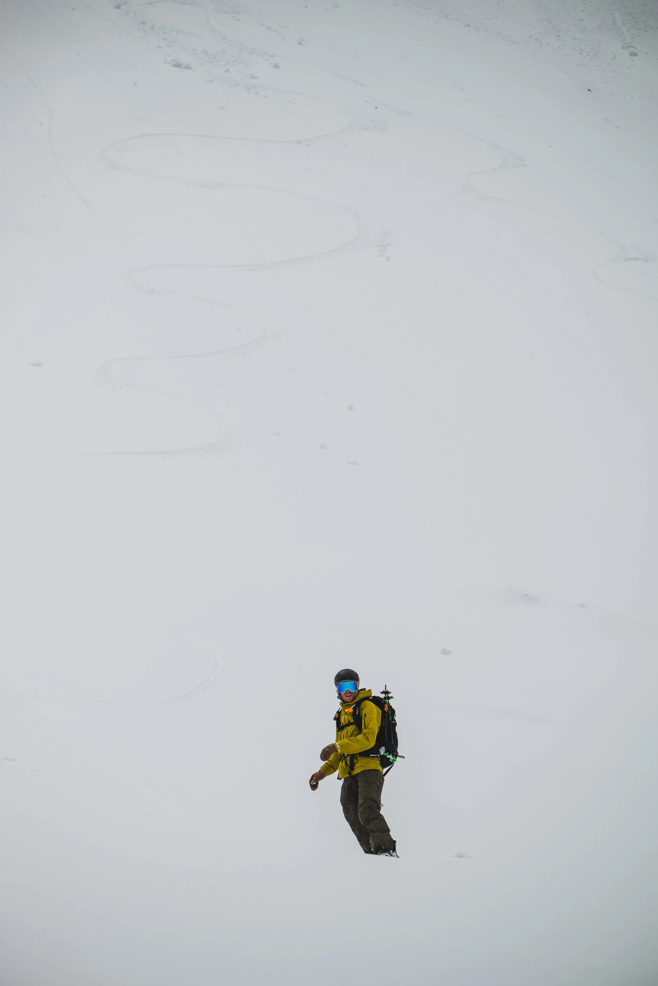 A person dressed in yellow and black winter gear, wearing a helmet and goggles, standing in deep snow with ski tracks in the background.