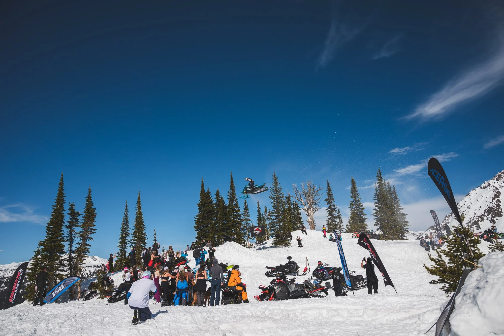 Snowy mountain scene with a skateboarder performing a jump in mid-air, surrounded by people, snowmobiles, and flags, under a clear blue sky with pine trees and snow-covered mountains in the background. Montana backcountry snowmobiling