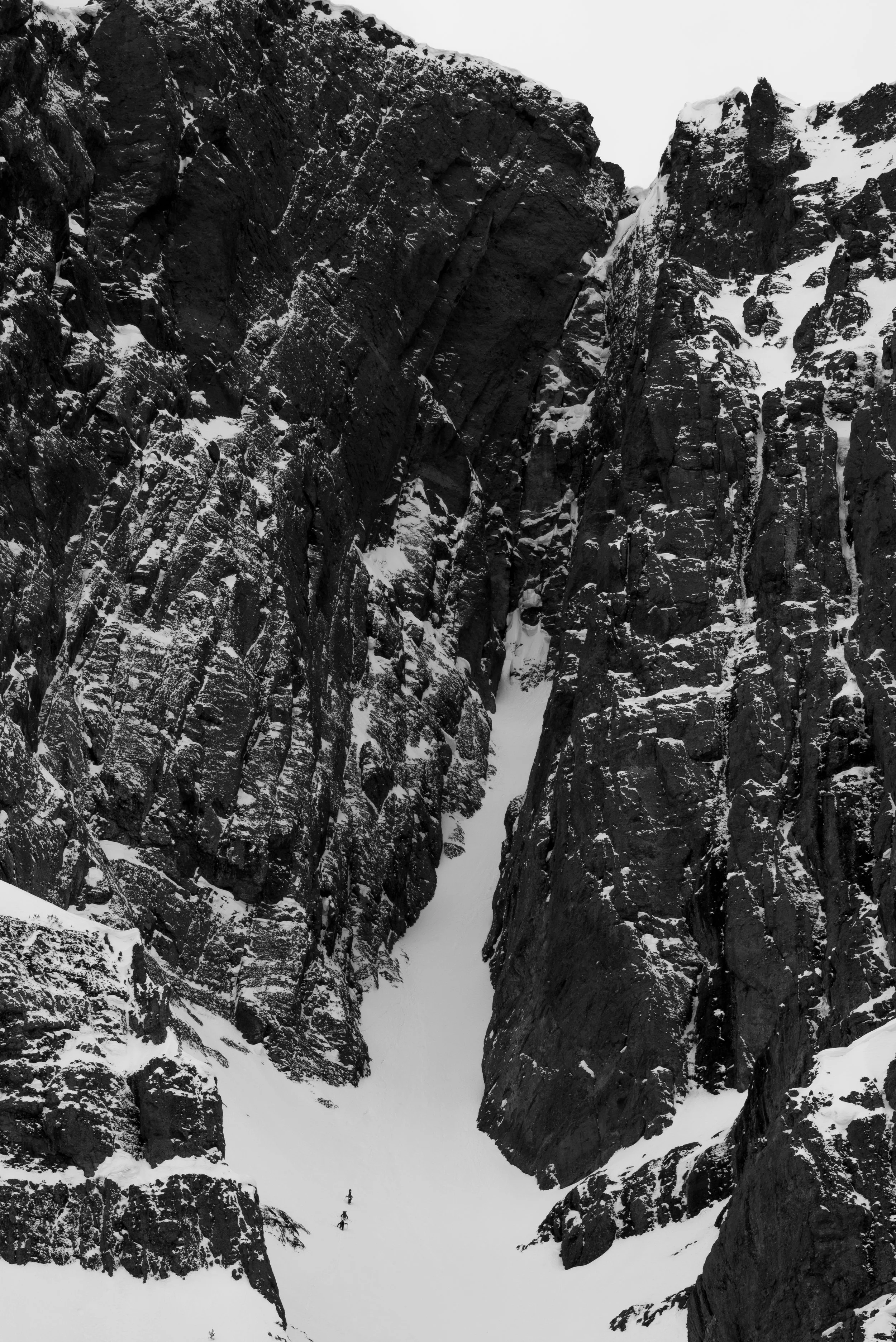 Snow-covered narrow canyon with high rocky walls and two tiny figures at the bottom, hiking or climbing.