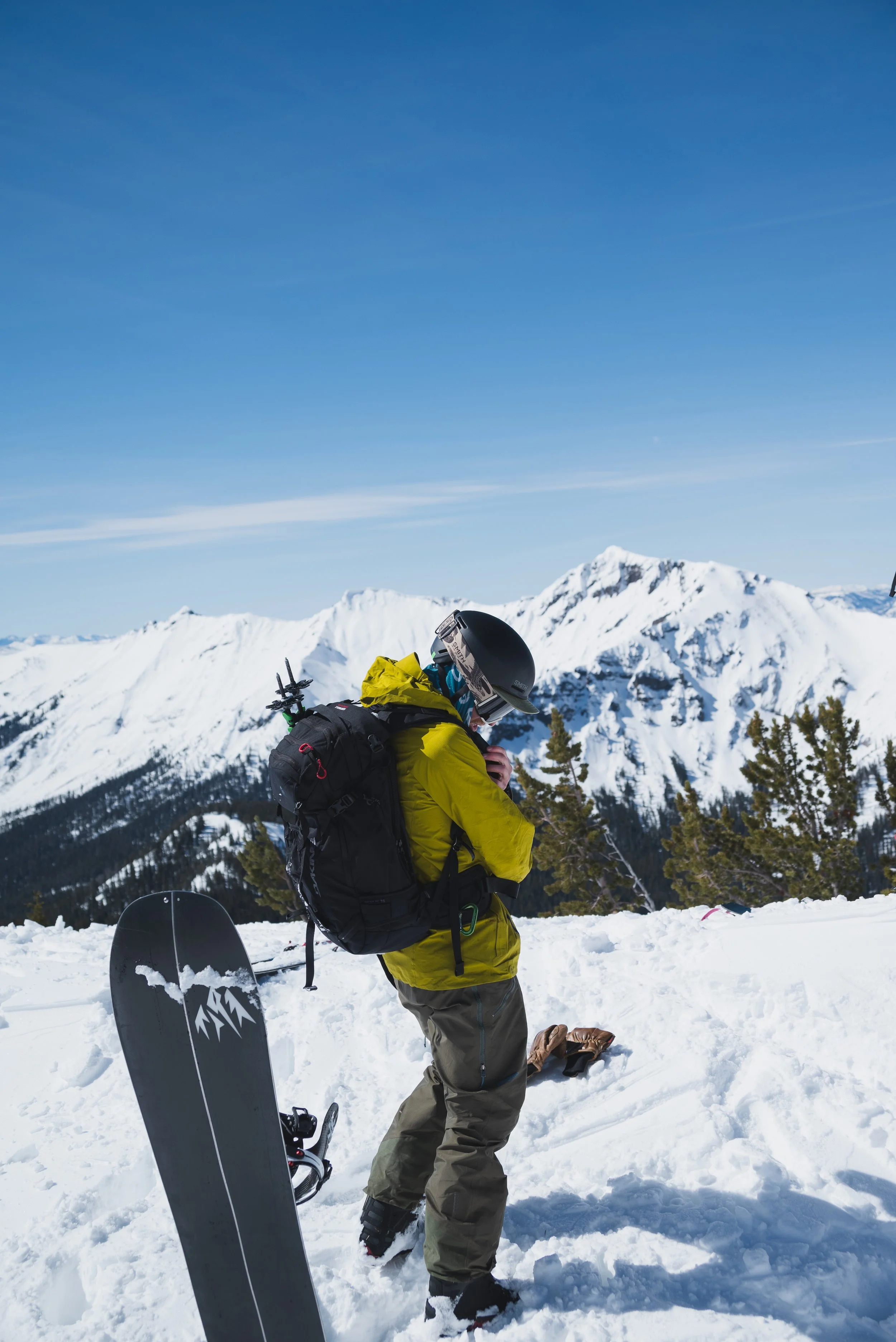 A person wearing a yellow snowboarding jacket, gray pants, and a black helmet standing in snowy mountains with snow-covered peaks and pine trees in the background, preparing for snowboarding.