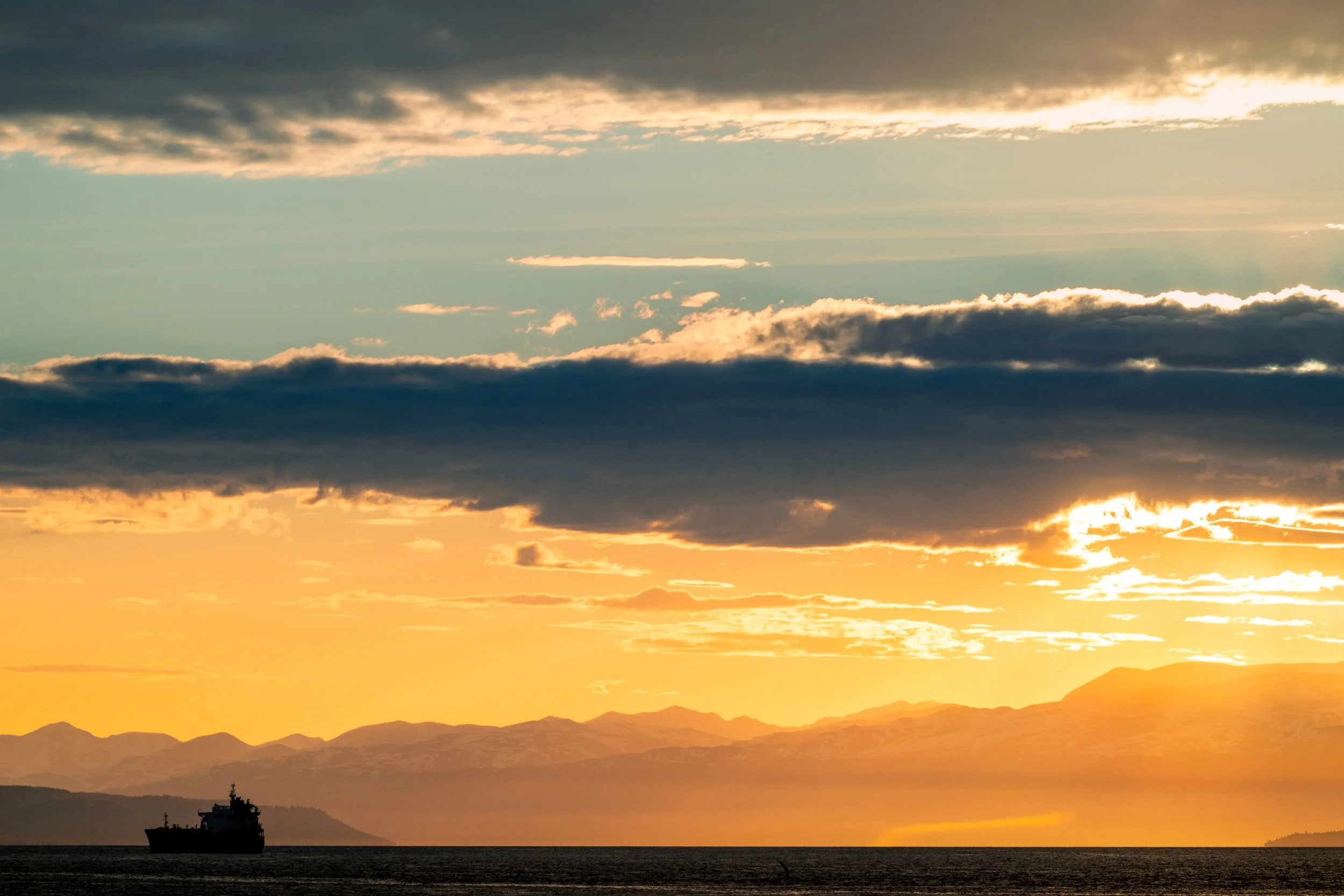 Sunset over ocean with dark clouds, distant mountains, and a ship sailing in the foreground.