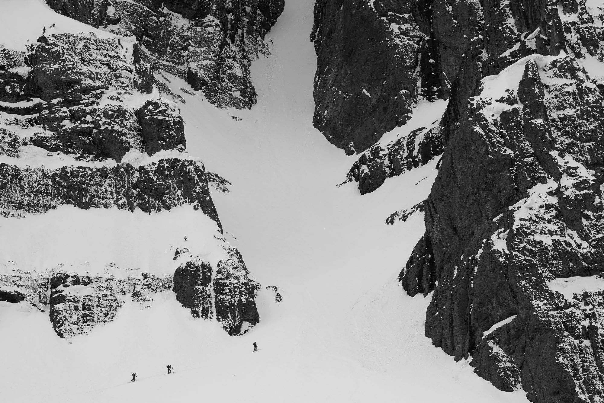 Three climbers ascending a snow-covered mountain between tall, dark, snow-dusted rocky cliffs. Montana backcountry skiing.