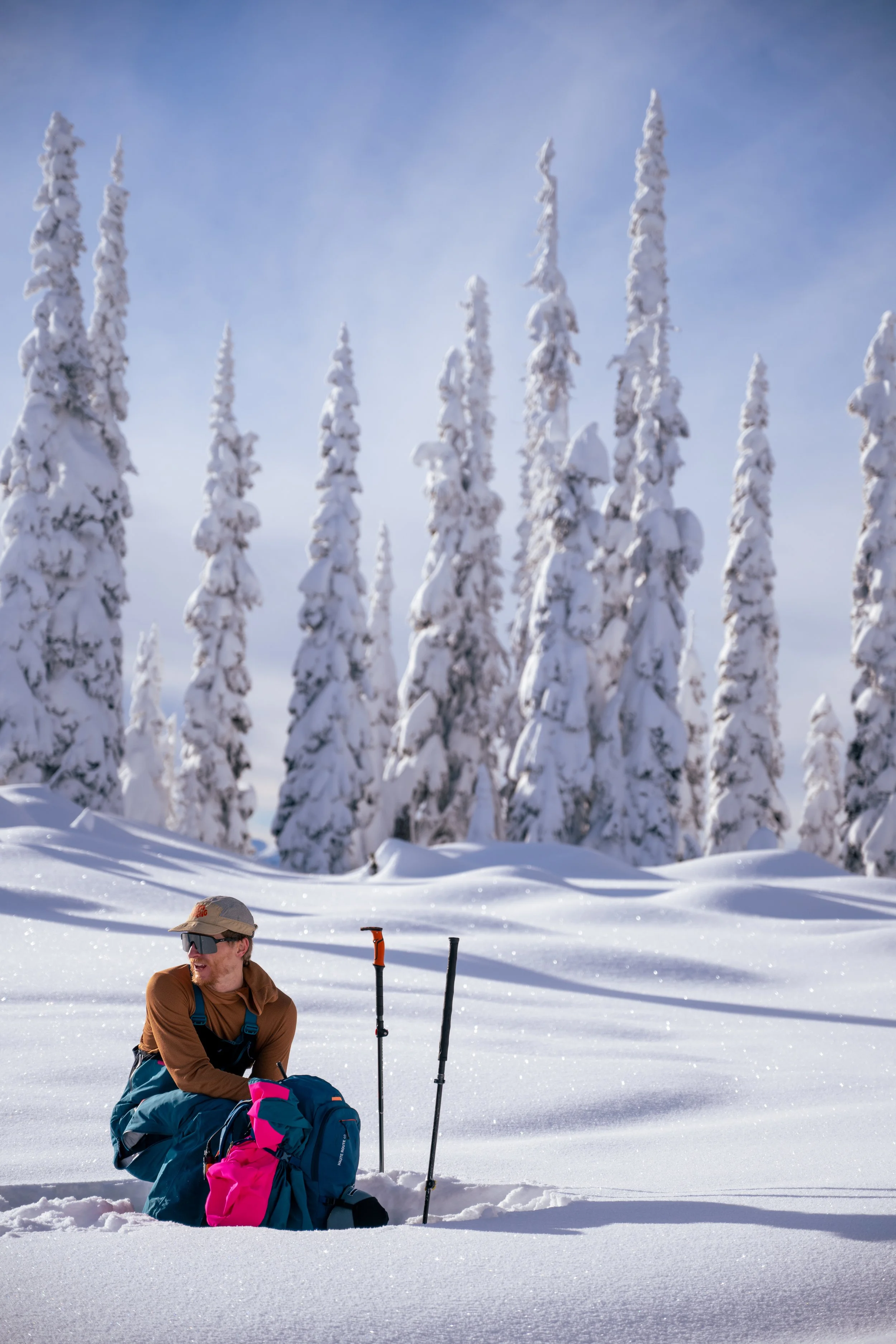 A person in outdoor winter gear sitting in deep snow with a backpack and trekking poles, surrounded by snow-covered trees under a blue sky.