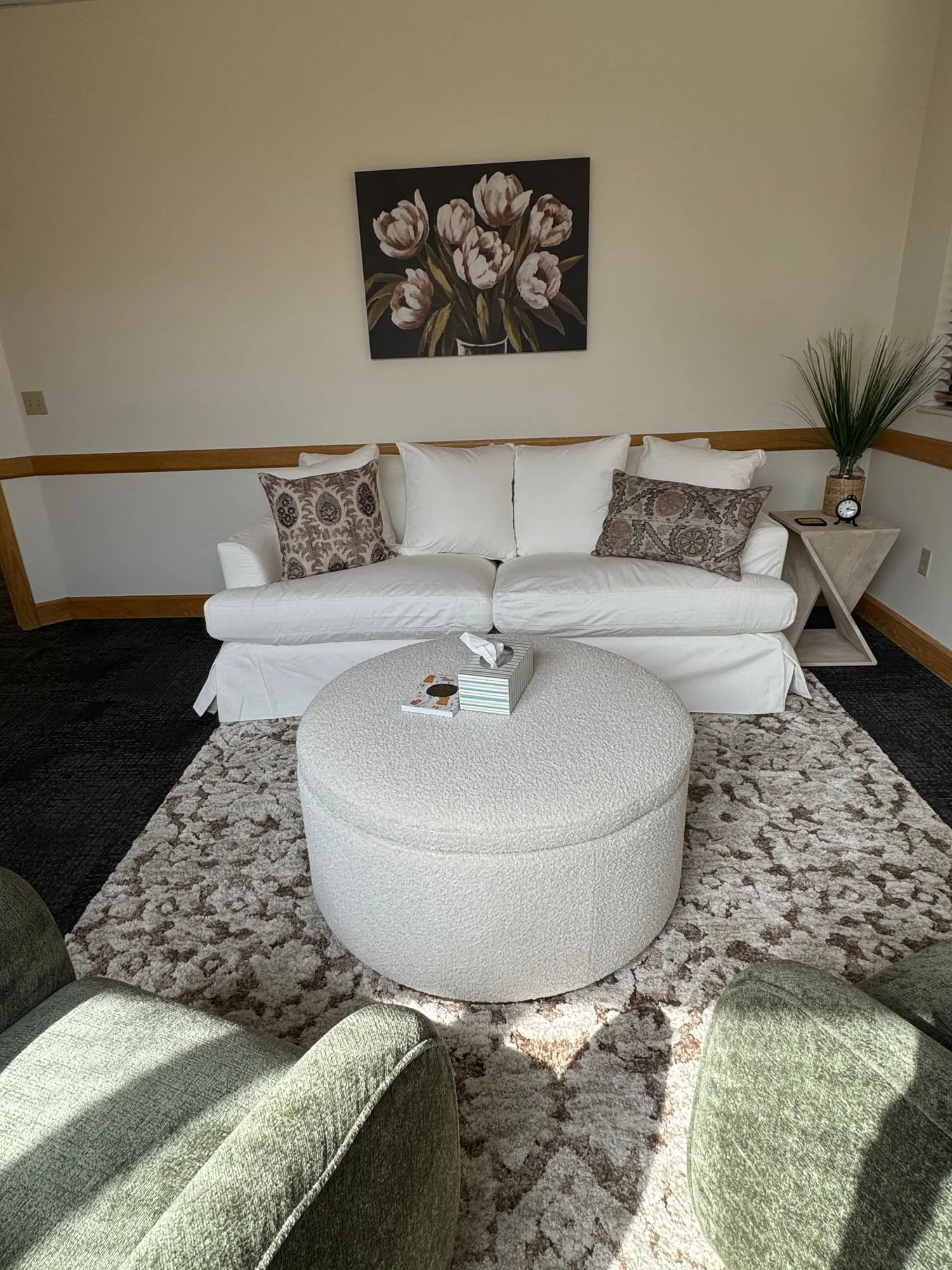 Modern living room with white sofa, patterned pillows, round ottoman, and floral wall art. Green chairs and decorative plant beside a framed painting. Neutral color scheme with a textured rug.