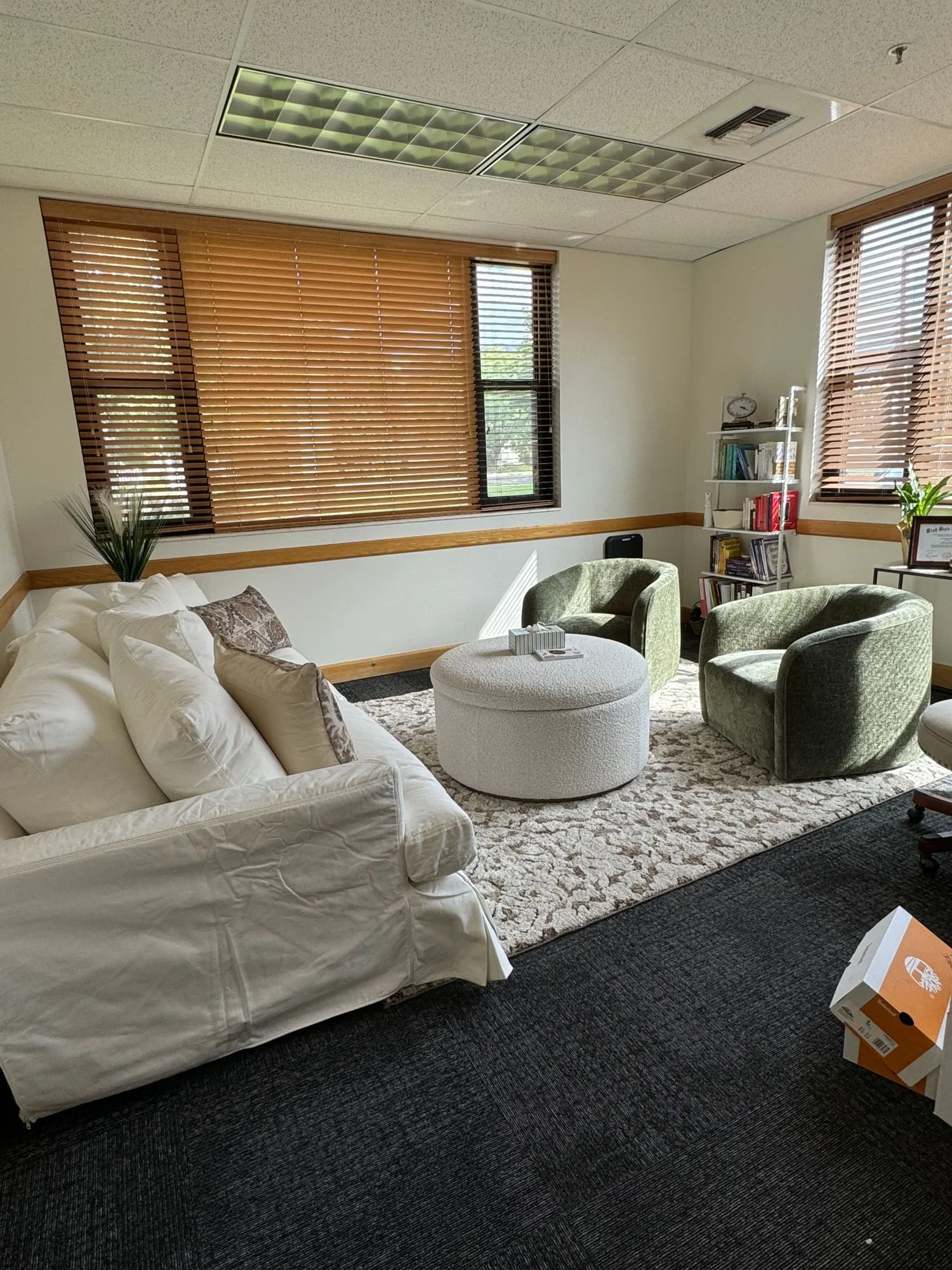 Cozy office seating area with a white couch, two green swivel chairs, a round ottoman, and a bookshelf, bathed in natural light from windows with wooden blinds.