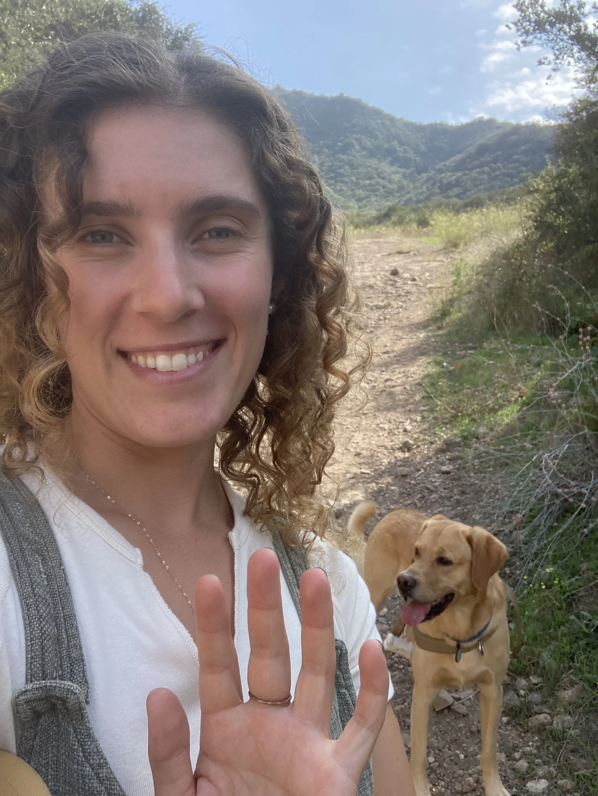 A woman with curly hair smiling at the camera while holding up her hand, wearing a white top and a backpack, with a yellow dog standing on a dirt trail in a mountainous, green landscape in the background.