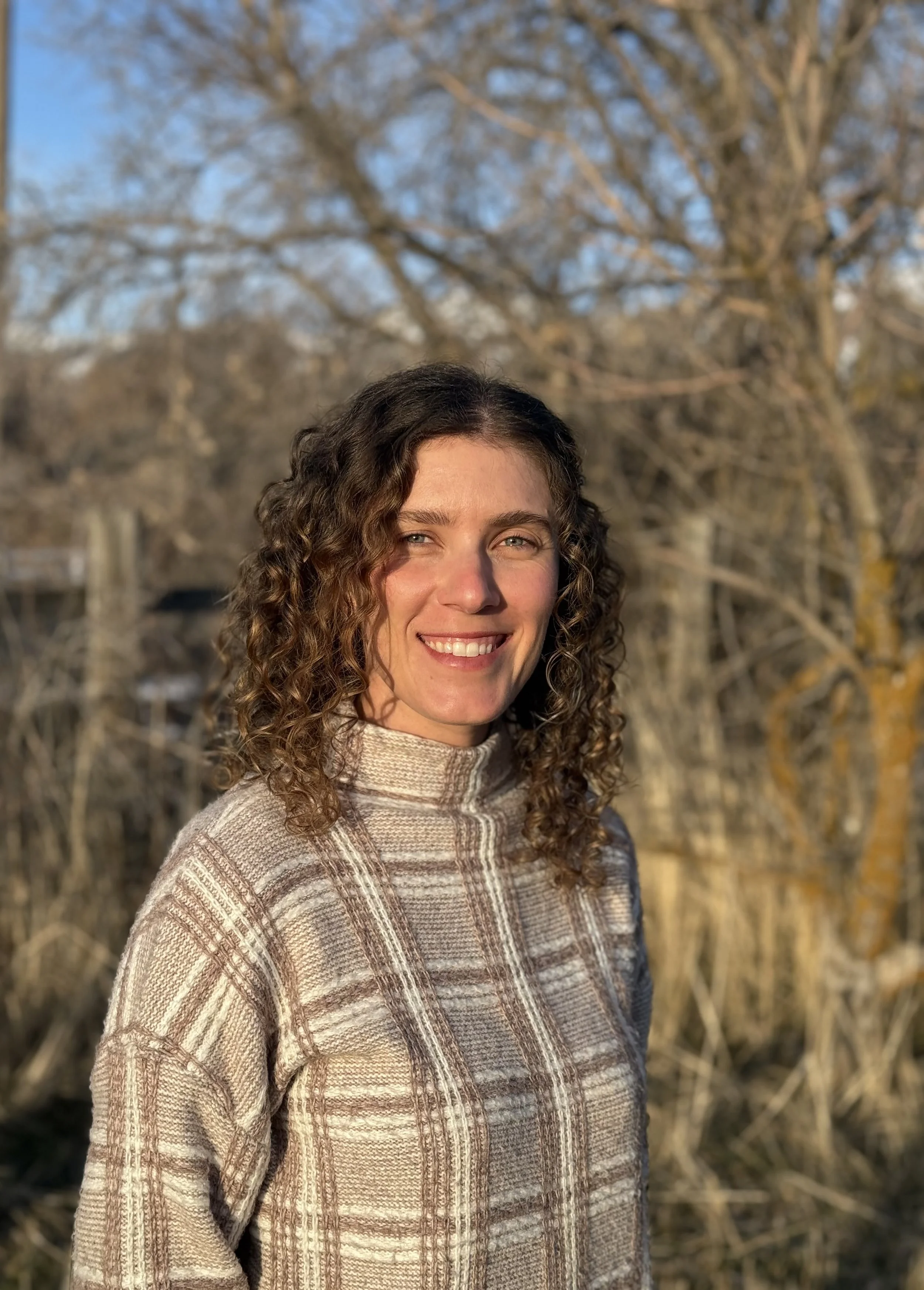 A woman with curly hair smiling outdoors during the daytime, wearing a patterned turtleneck sweater, with trees and a blue sky in the background.
