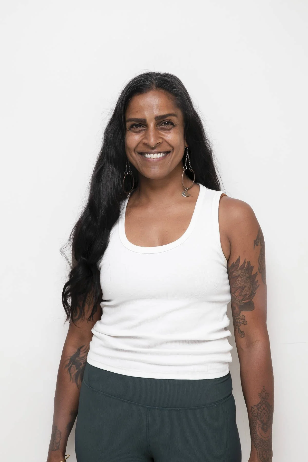 Smiling woman with long black hair and tattoos, indoors with wooden floors and bright natural light.