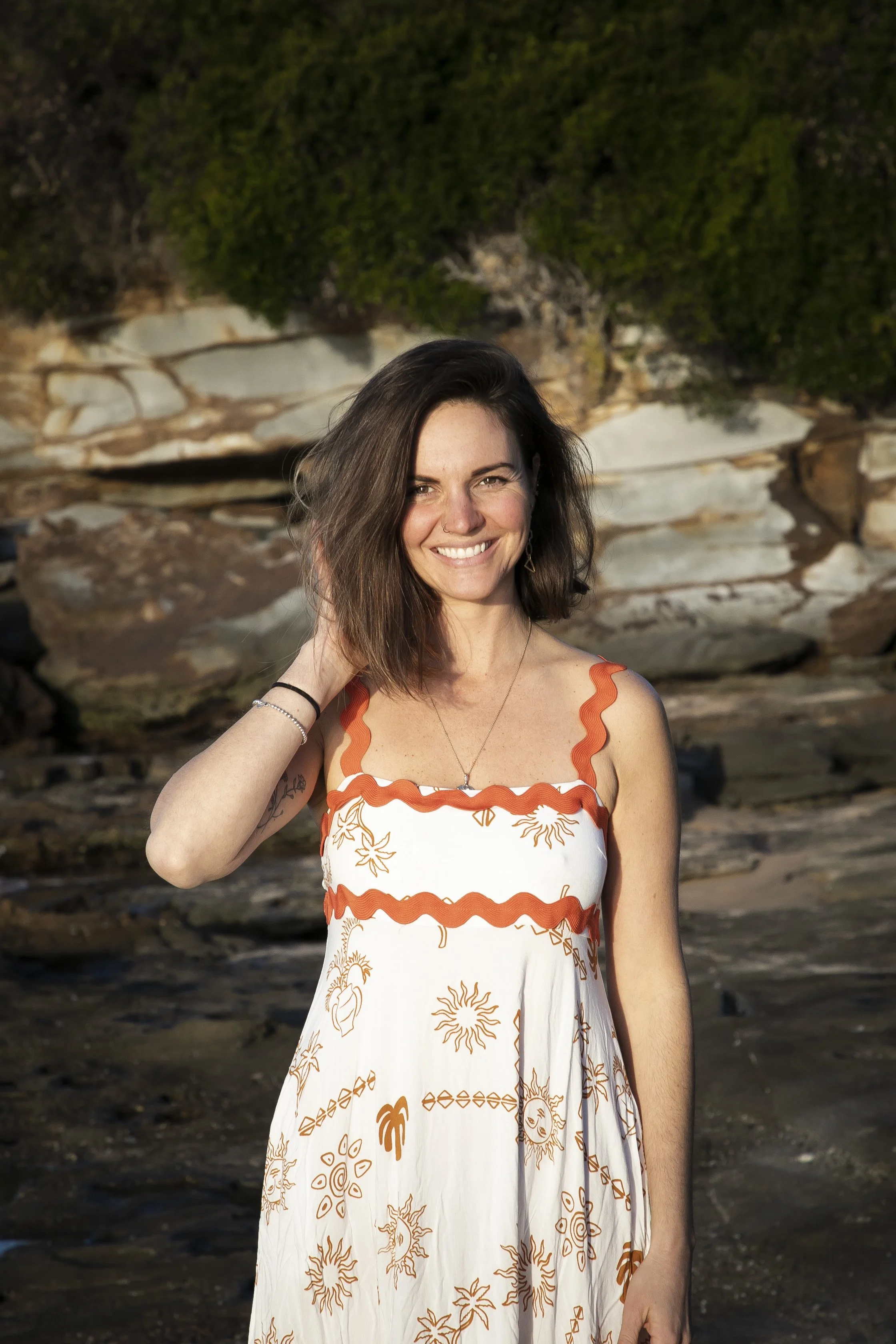 Woman smiling outdoors, wearing a white sundress with orange patterns, standing in front of rocky terrain.