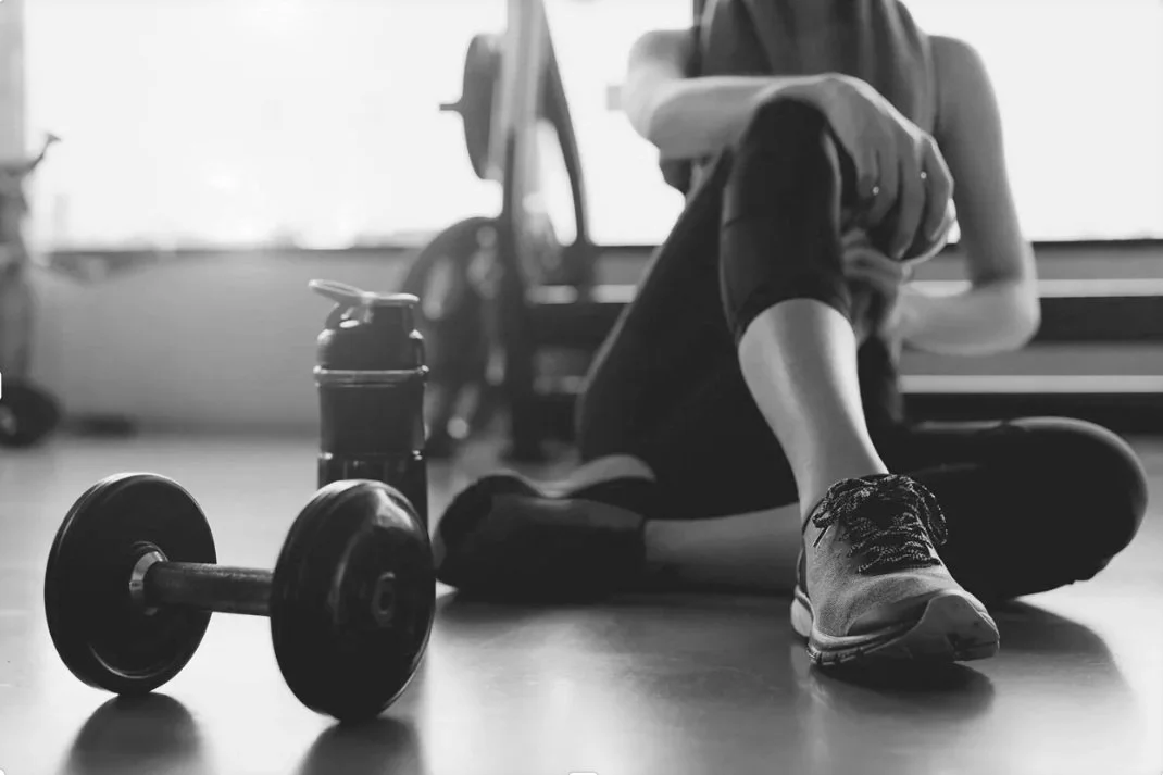 A reflective black and white scene at 901fitlife Memphis with an open workout logbook next to a '901' engraved dumbbell, a '901fitlife' etched water bottle, and a resting member, with a distinct red and green neon sign in the deep background