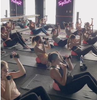 A group fitness class of women in activewear performing core stability and upper body dumbbell exercises using Pilates balls on mats. A large, glowing neon sign on a brick wall reads 'Better Together', conveying a supportive community workout atmosph