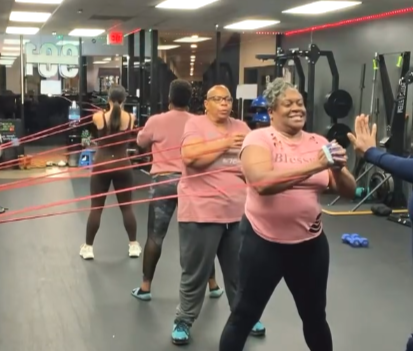 A group of people exercising at a gym, with one woman in the foreground smiling and giving a high five to another person, while others are using resistance bands and weights in the background.