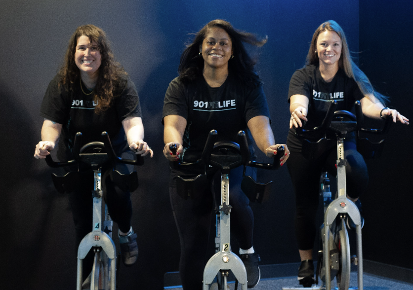 Three smiling women in branded 901FitLife shirts participating in an indoor cycling spin class in Memphis, highlighting the gym's supportive community.