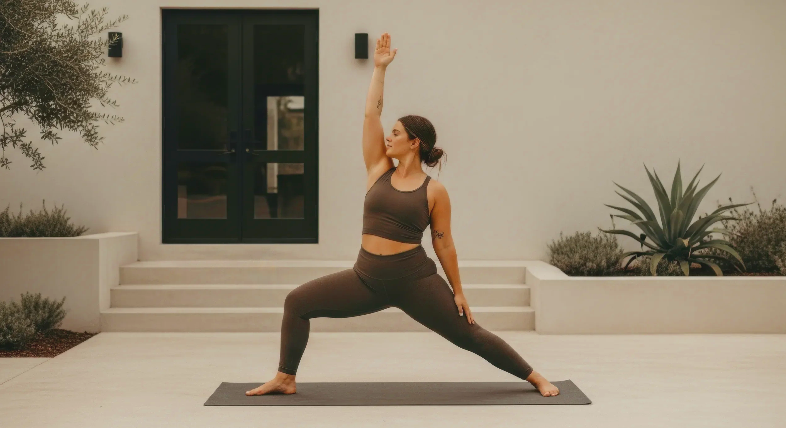 A woman practicing yoga outdoors on a black mat, standing in a warrior pose with one arm raised and the other hand on her thigh, in front of a modern house with white steps and plants.