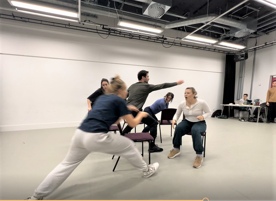 Group of people in a rehearsal or acting workshop, performing dynamic movements in a room with chairs and a table in the background.
