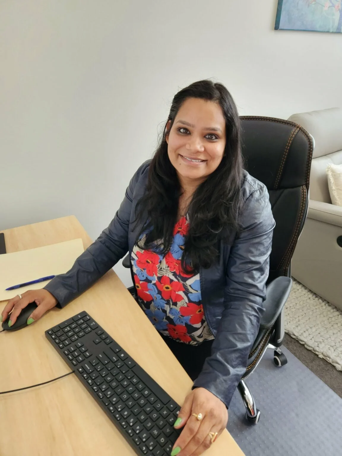 Woman sitting at desk with computer keyboard, mouse, notebook, and pen, smiling at camera in an office.
