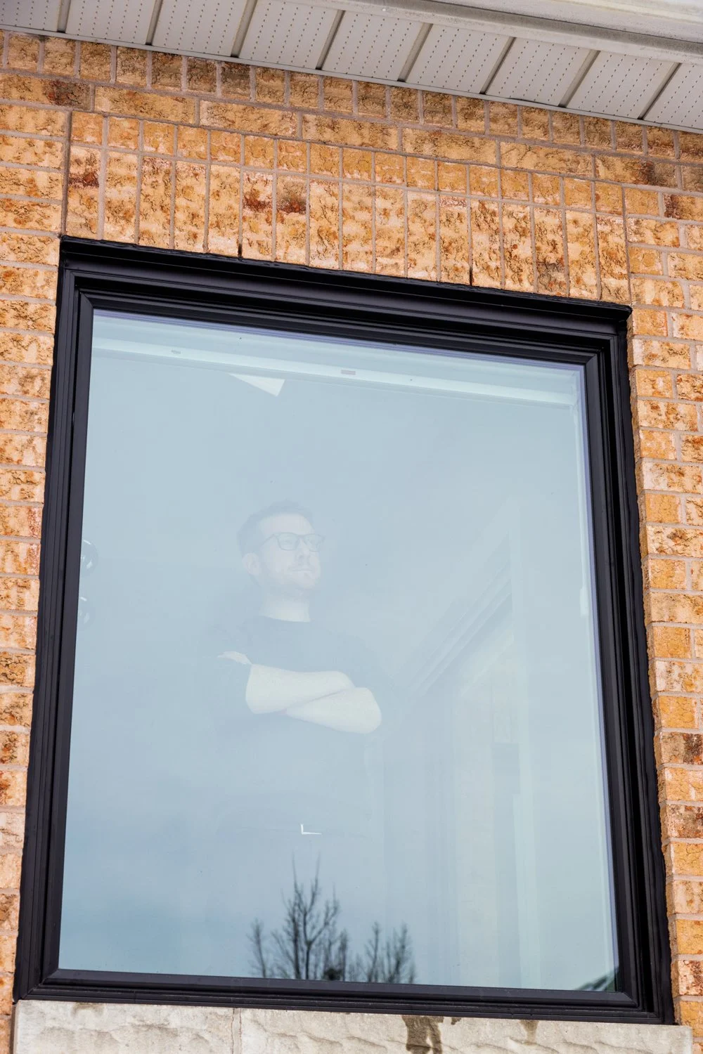A man wearing glasses and a black t-shirt looking out a large window with a black frame, reflecting a cloudy sky and leafless trees outside, set in a brick building.