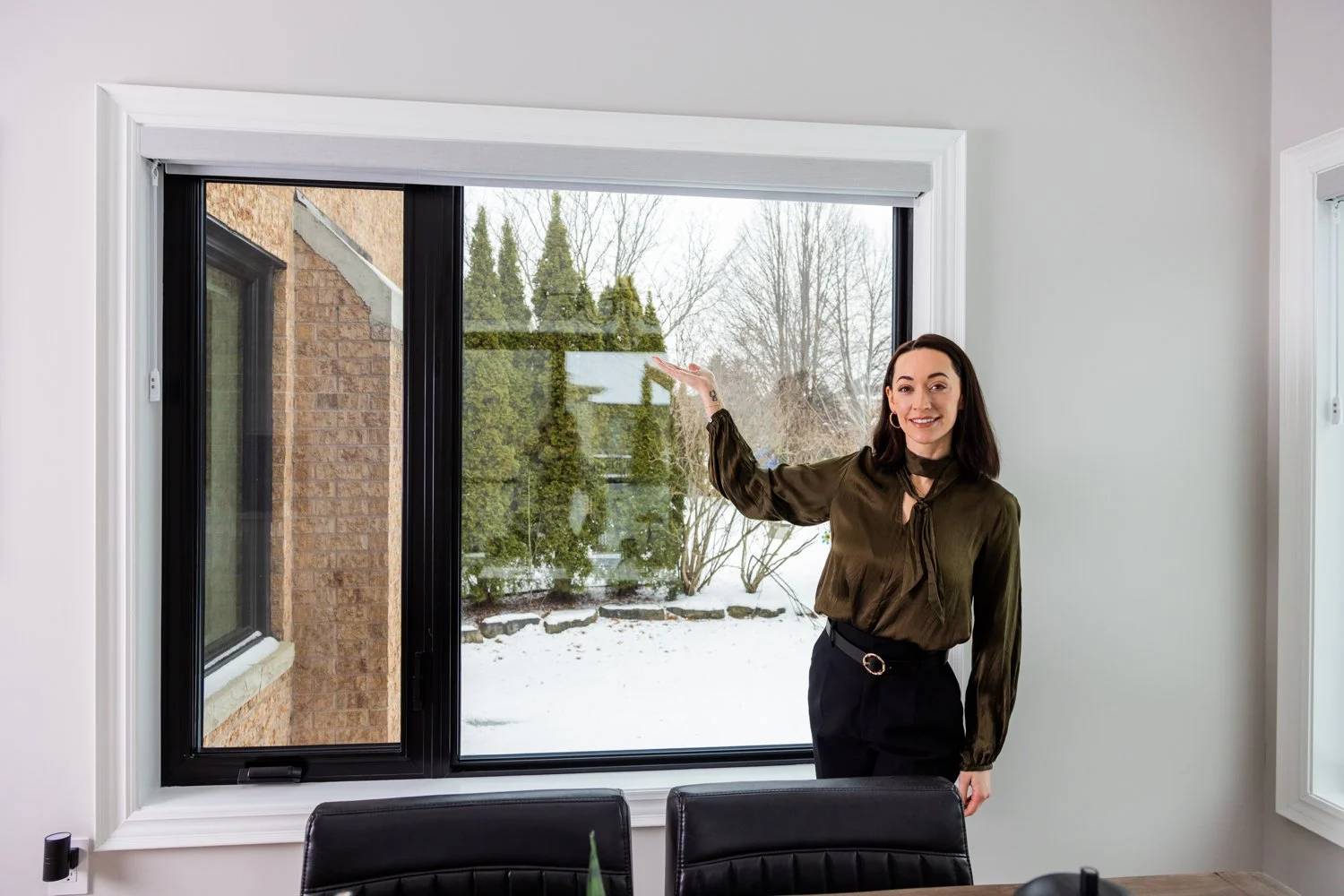 Woman in a green blouse standing inside, pointing at a large window showing a snowy outdoors scene with trees and a brick building.