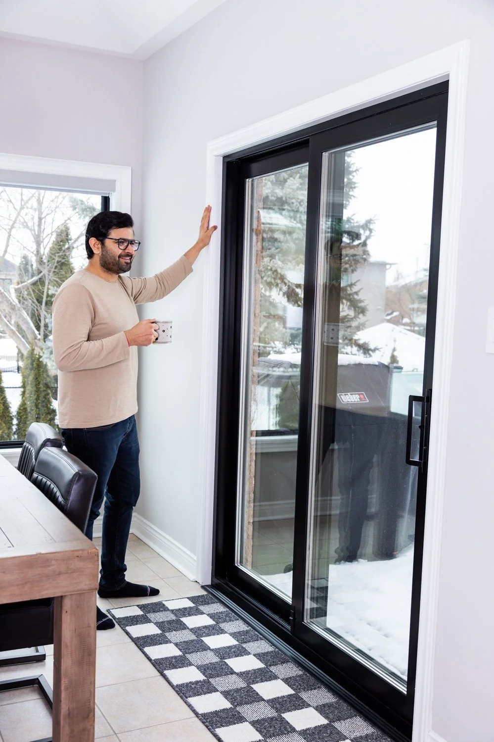 A man with glasses standing inside near a sliding glass door, holding a mug, and looking outside at a snowy yard with trees.