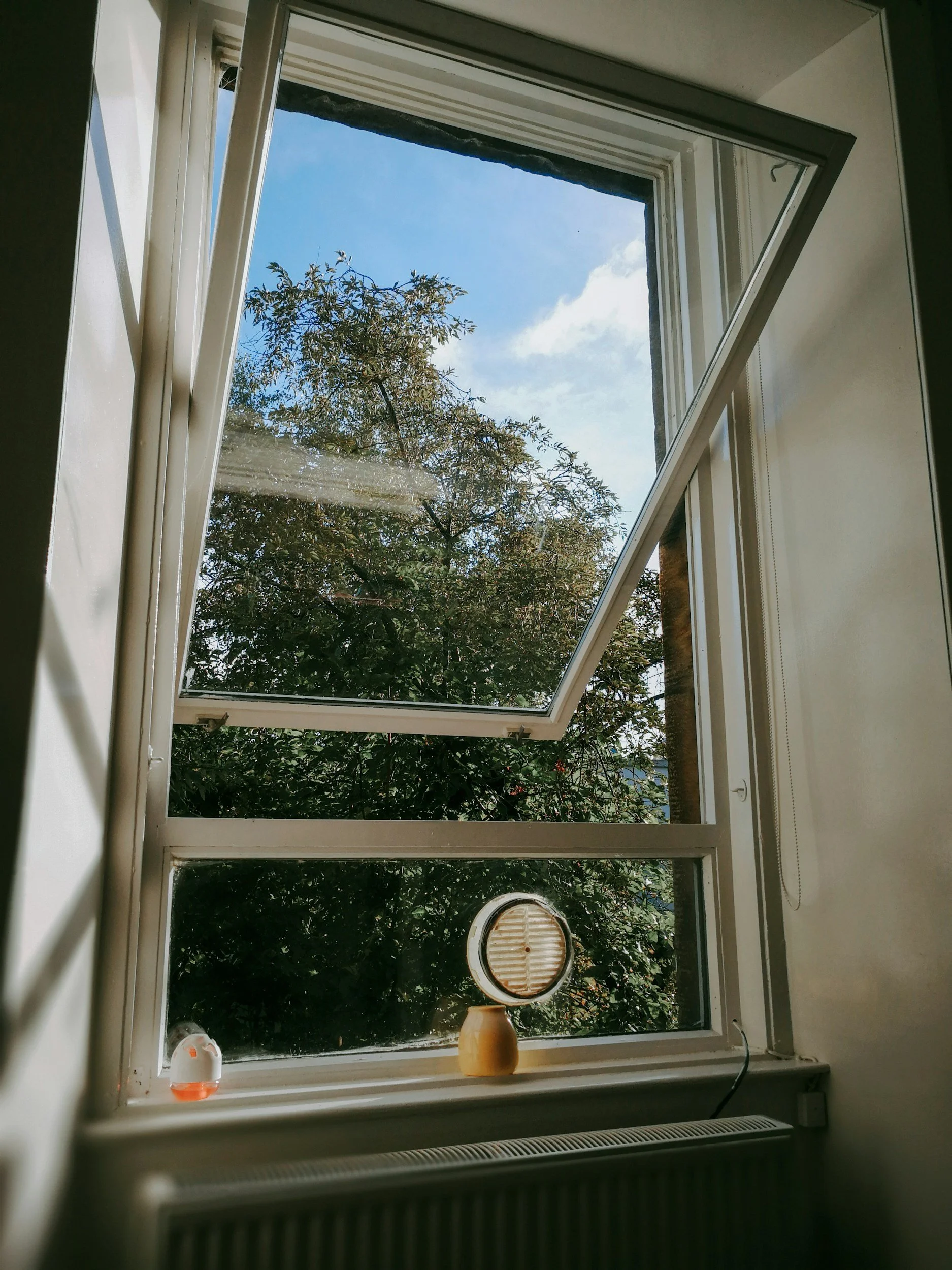 Open window overlooking a tree and blue sky with some clouds, with decorative items on the windowsill including a small humidifier and a round mirror.
