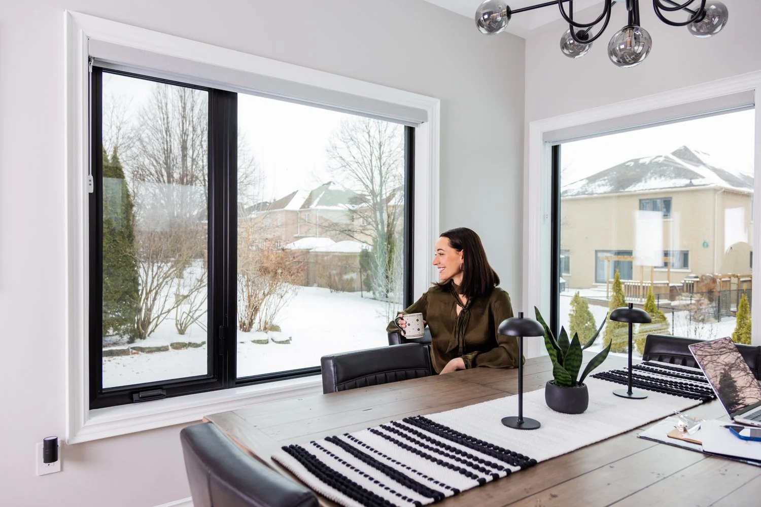 A woman sitting at a dining table in a bright room, looking out a large window at a snowy outdoor scene, holding a mug and smiling.