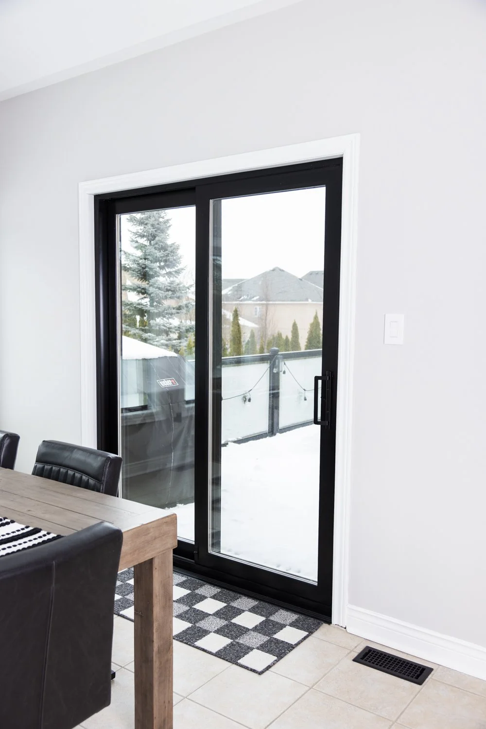 A sliding glass door leading to a snow-covered backyard with trees and houses in the background, part of a modern dining room with a wooden table and black chairs.