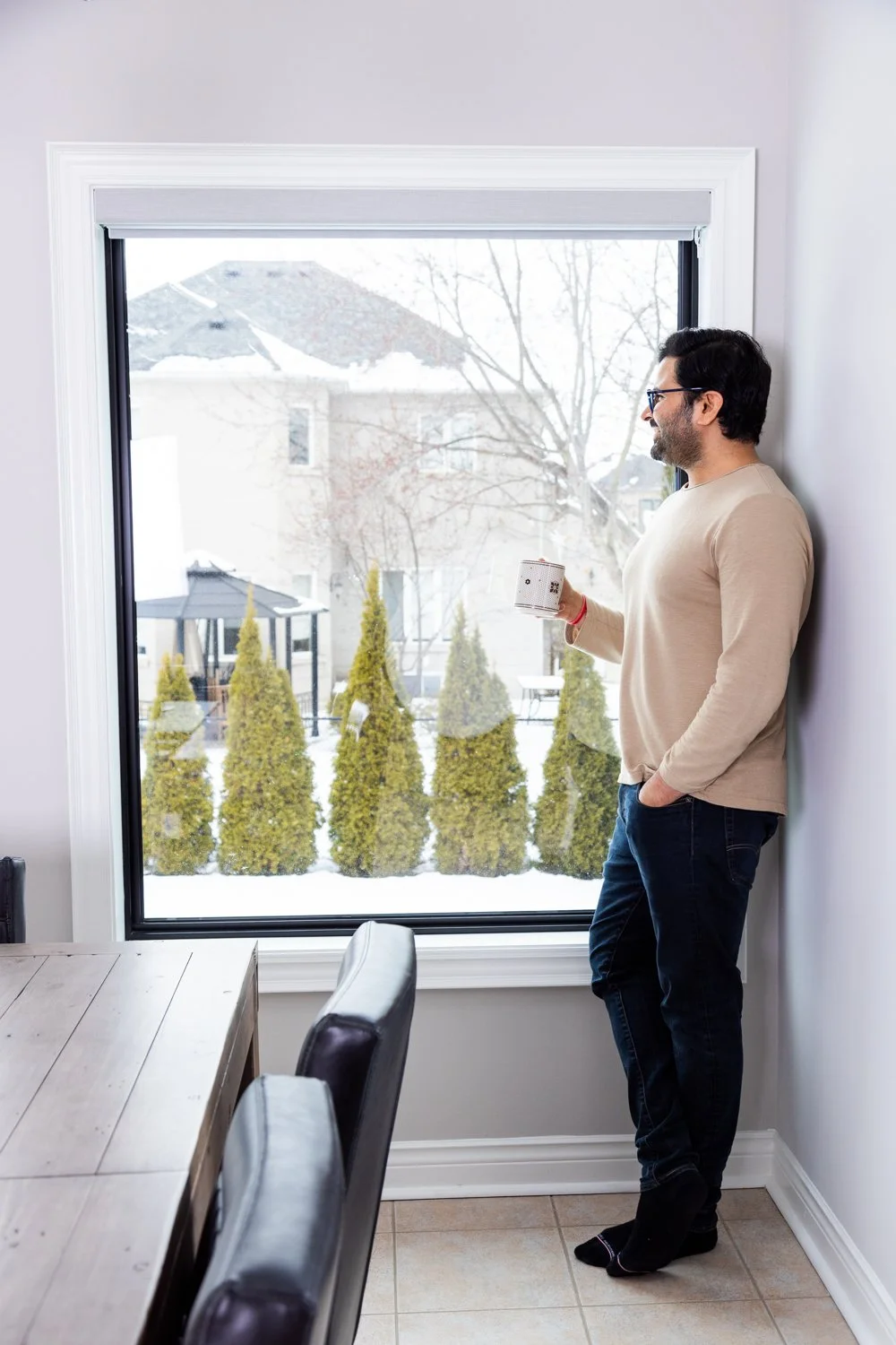 A man in a beige sweater and dark jeans stands by a window indoors, holding a mug and looking outside at snow-covered trees and a house with a damaged roof.