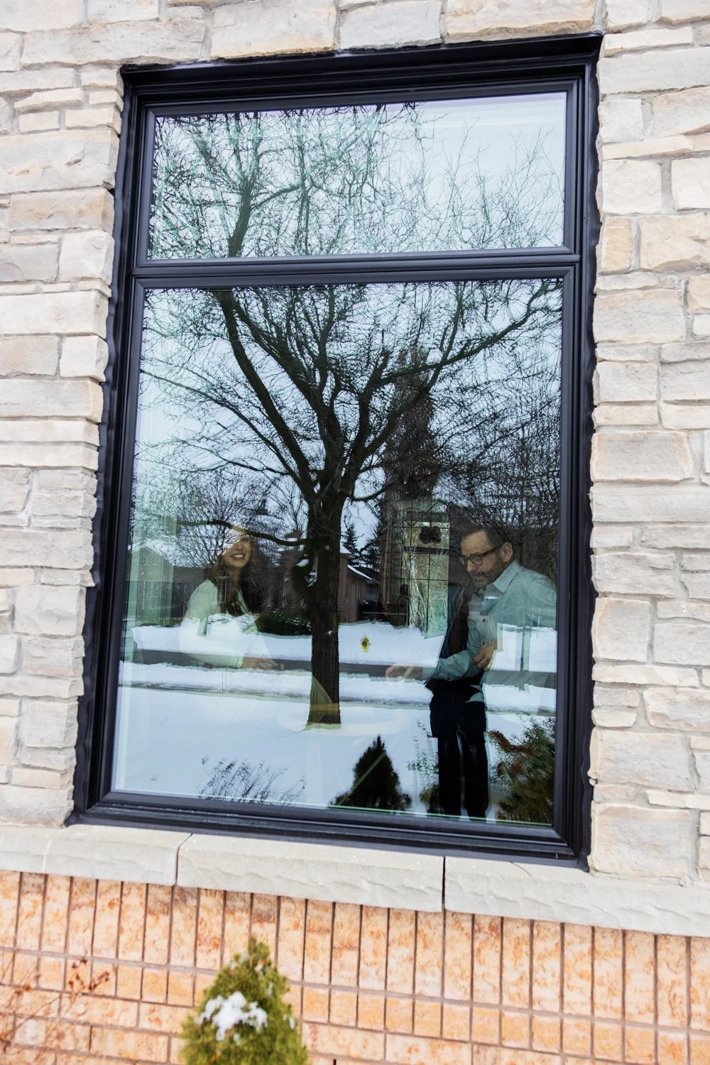 Reflected image of two people smiling and looking at a device, seen through a window, with a snow-covered yard and a leafless tree outside.
