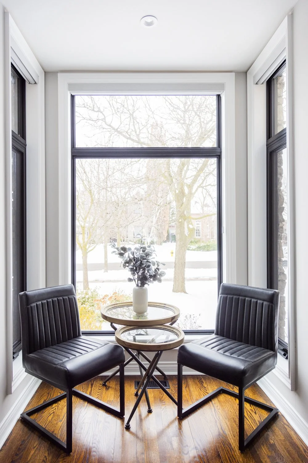 A cozy corner with two black leather chairs, a small round wooden table with a glass top, and a white vase with greenery, set in a sunlit breakfast nook with large windows and a view of outdoor trees and snow.