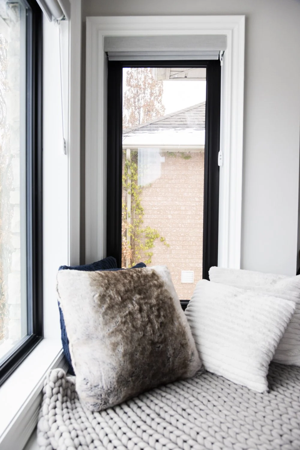 Interior view of a window seating nook with pillows, looking outside to a brick house and trees.
