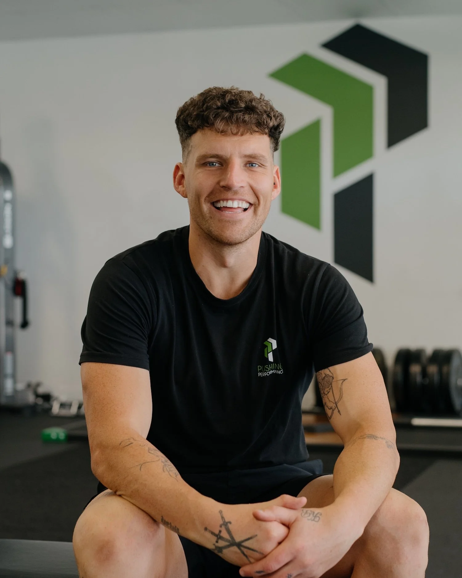 Man sitting in a gym, smiling, with tattoos on his arms, wearing a black t-shirt with 'Pushing Performance' logo.